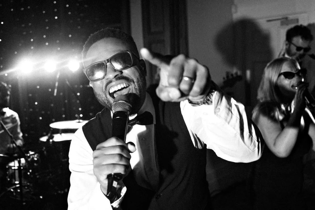 Black-and-white concert scene: smiling singer in sunglasses and tuxedo points at the camera while gripping a microphone; band performs behind him.
