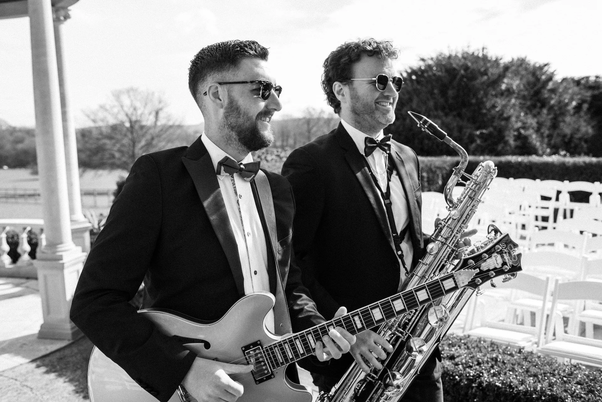 Two men in tuxedos and sunglasses smile outdoors, one holding a guitar, the other a saxophone, standing near rows of white chairs in daylight.