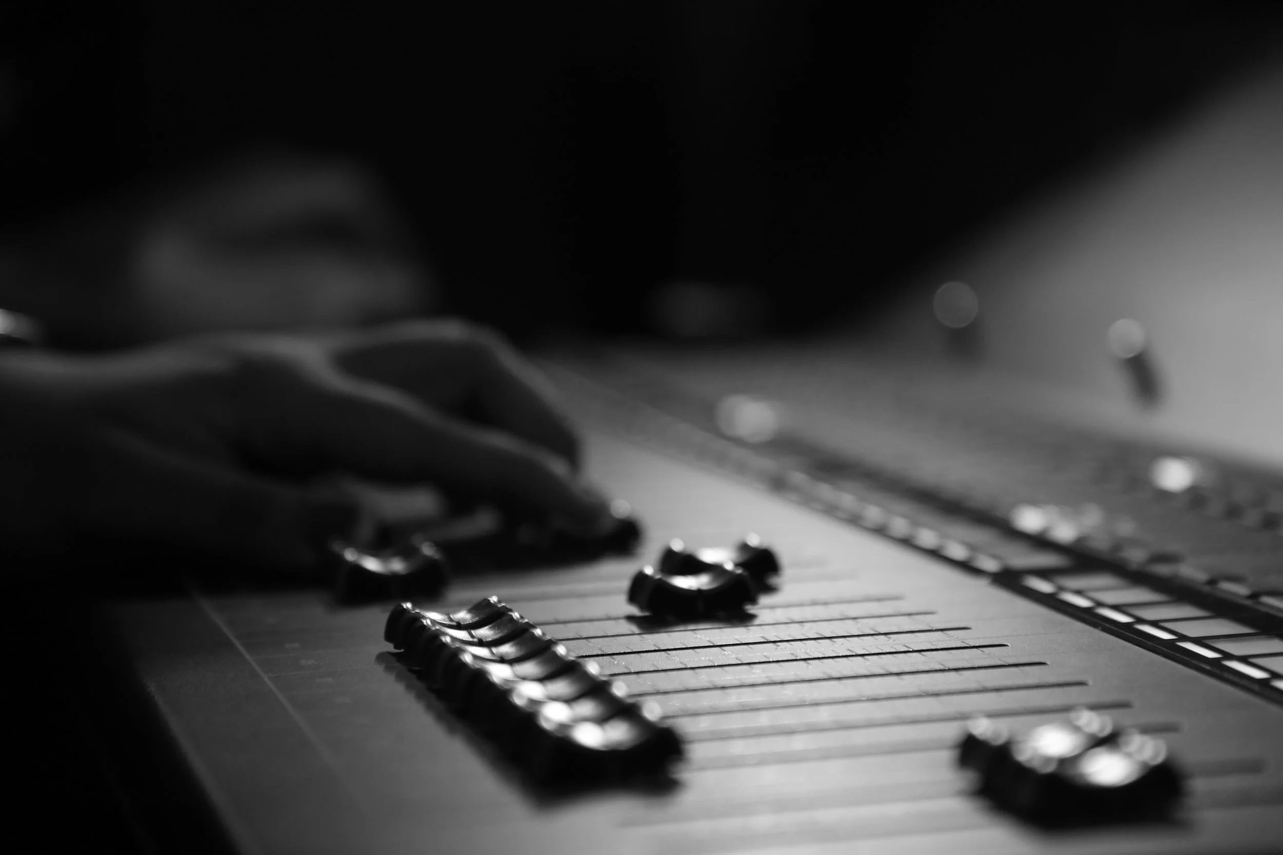 A close-up of a hand sliding controls on a sound mixer, with shiny knobs in focus and the rest fading into soft darkness.