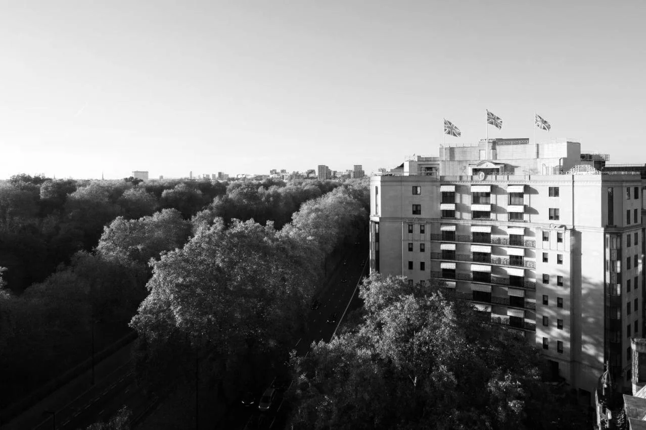 Black-and-white view of The Dorchester beside Hyde Park, its elegant facade with flags on the roof overlooking tree-lined parkland and London’s skyline.