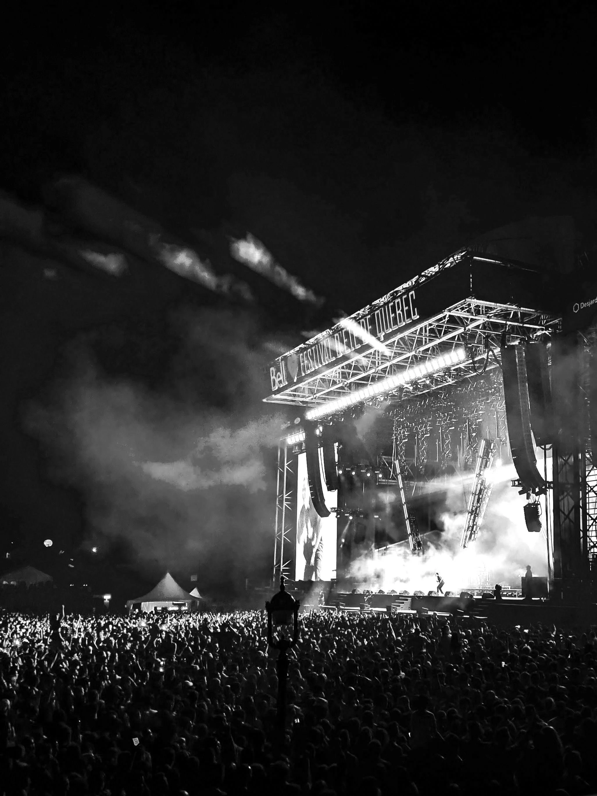 A vast crowd faces a towering outdoor stage, with smoke drifting, bright lights blazing, and a performer silhouetted against screens and rigging.