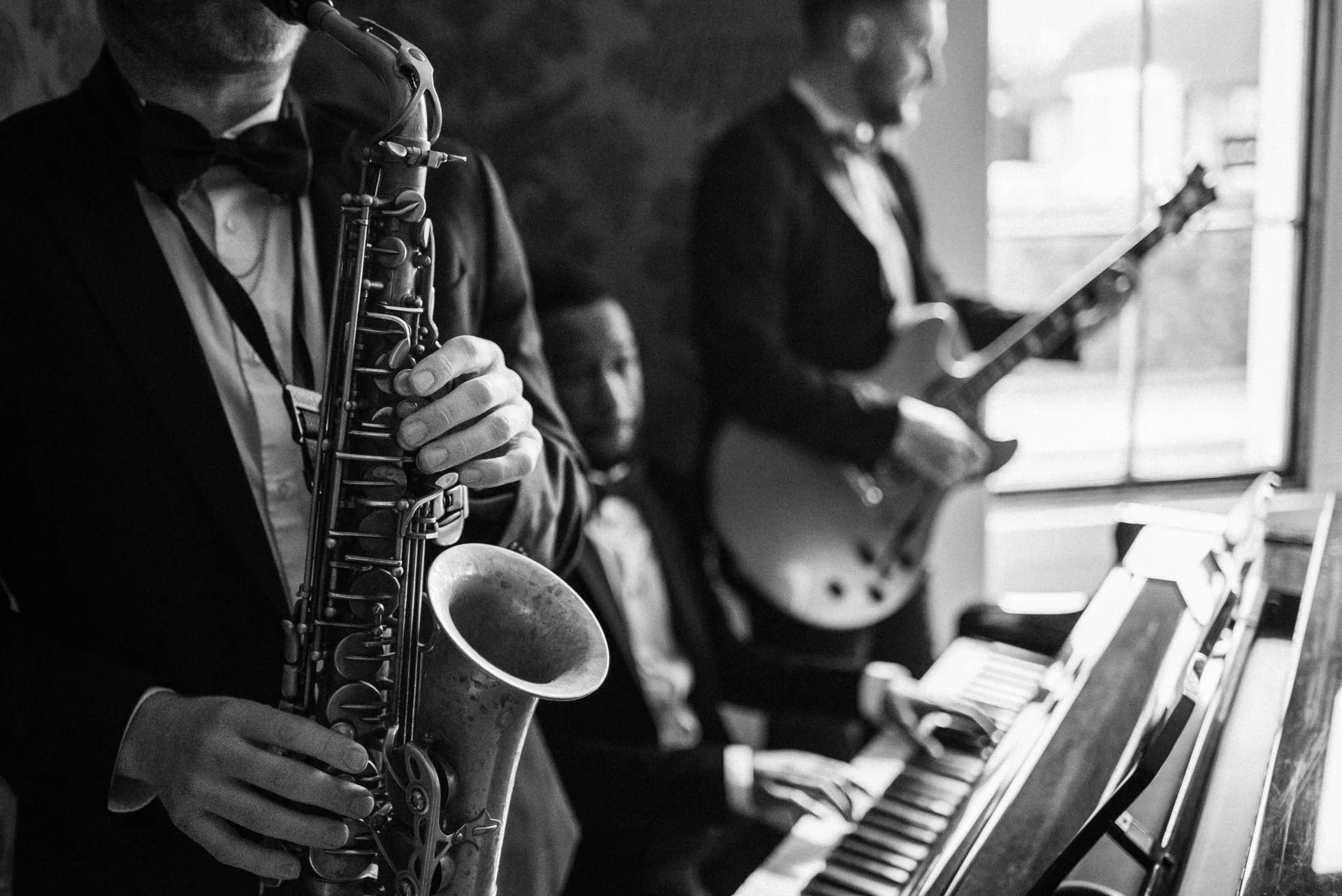 A saxophonist in a tux stands beside a pianist at a grand piano, while a guitarist plays near a bright window in the background.