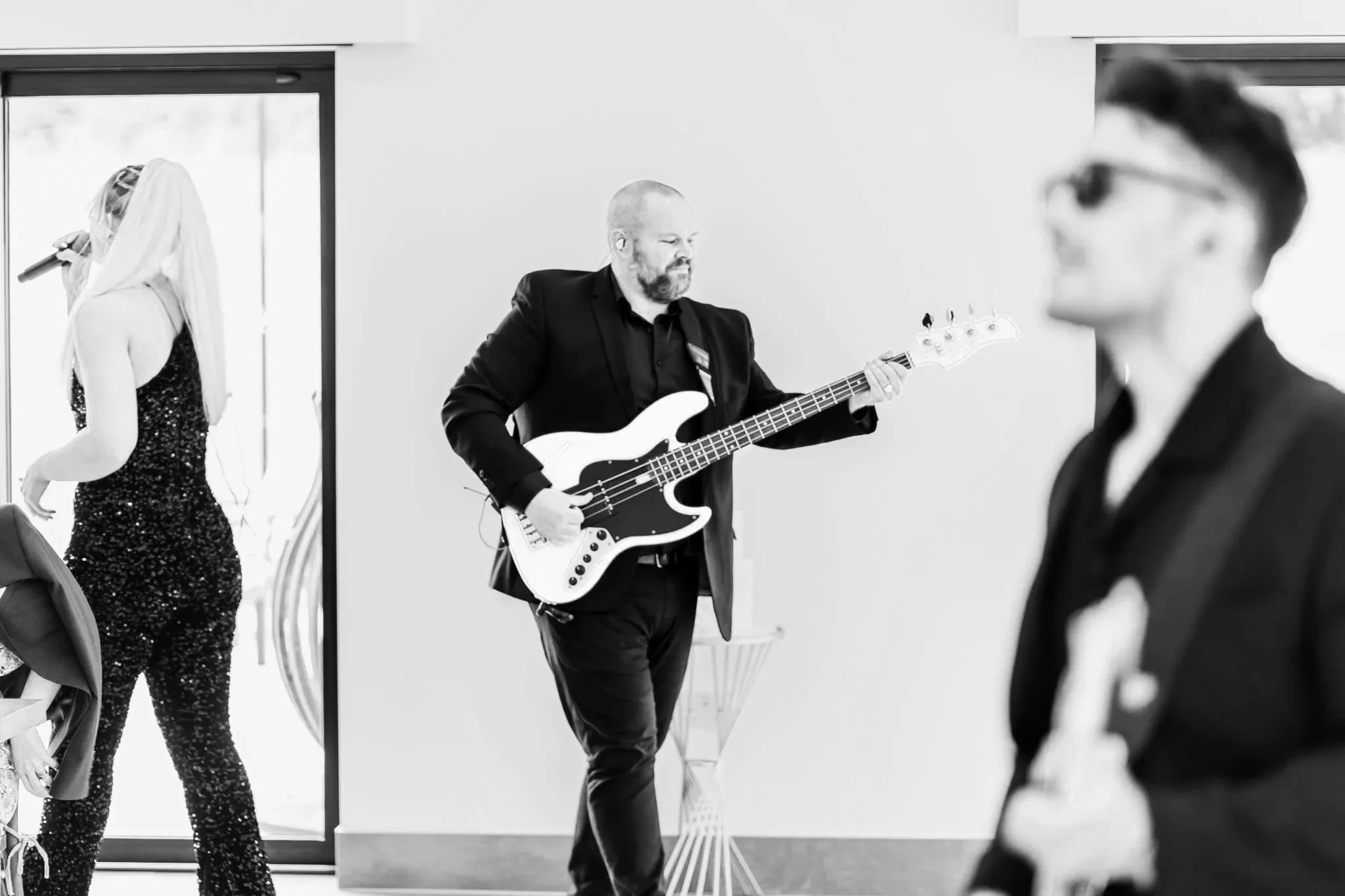 A bassist in a black suit plays a bright electric bass while a sequined singer performs nearby and another guitarist stands blurred in front.