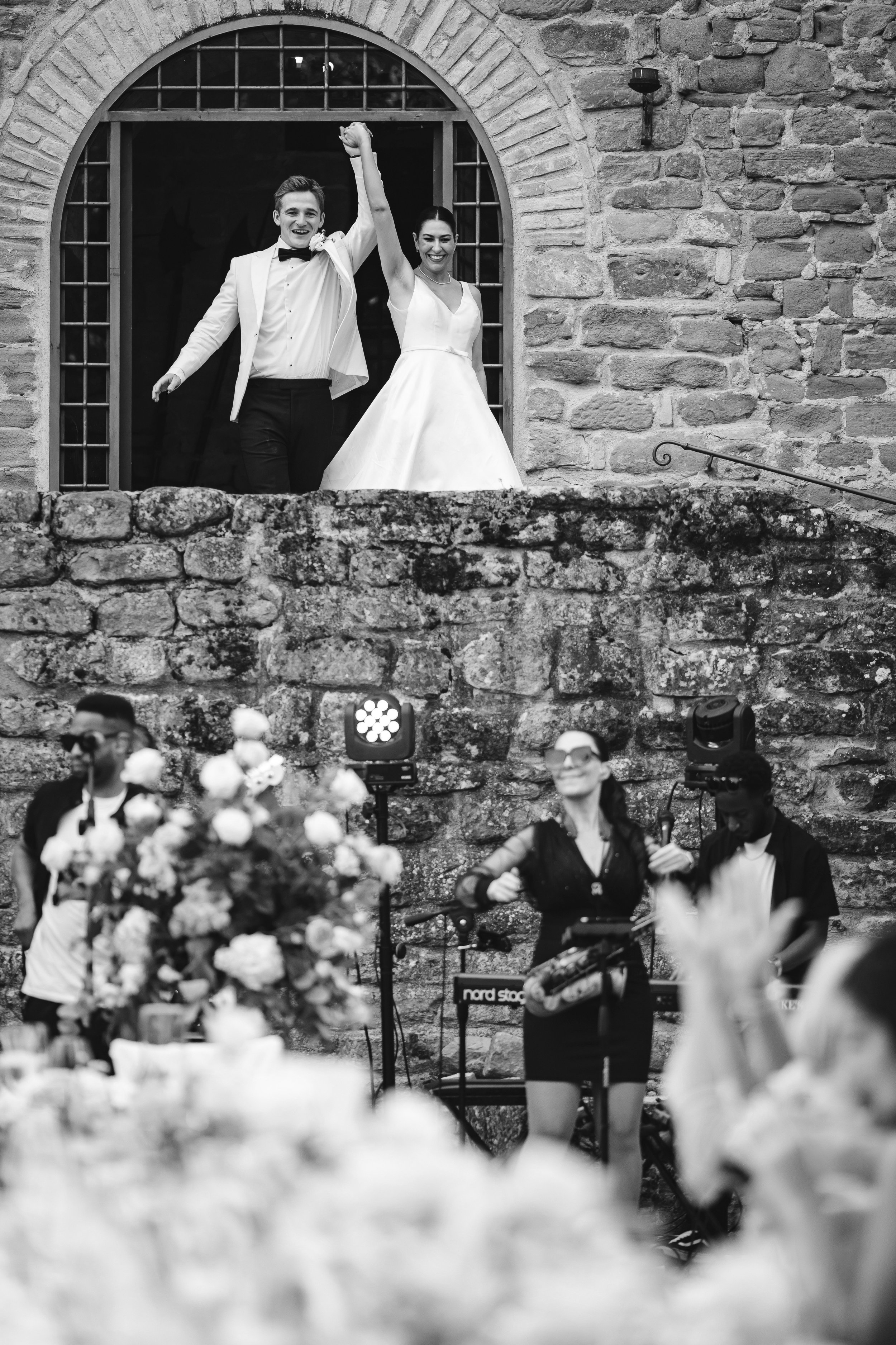 A bride and groom stand in a stone archway, smiling and raising joined hands in triumph, while musicians perform below amid flowers and cheering guests.