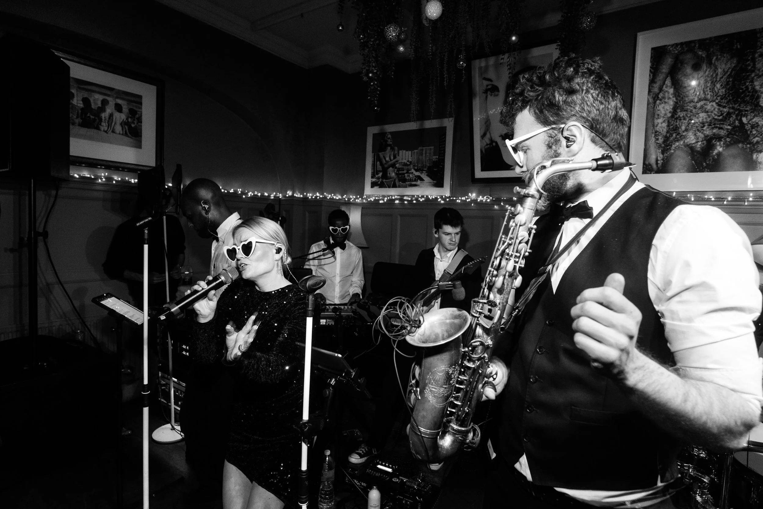 Black-and-white photo of a live band: a singer in heart-shaped sunglasses sings into a mic as a saxophonist plays beside her; band behind them.