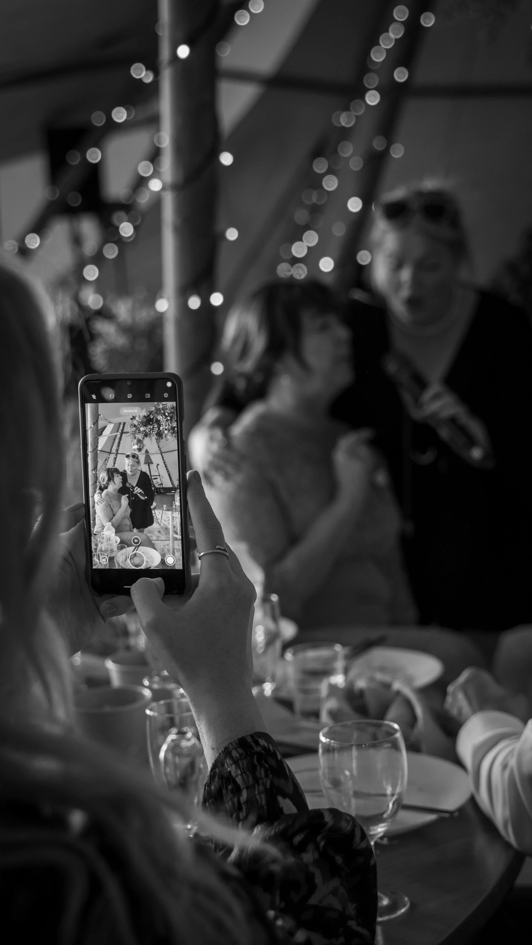 A guest holds up a phone, photographing two smiling women embracing at a candlelit table, with soft fairy lights glowing behind them.