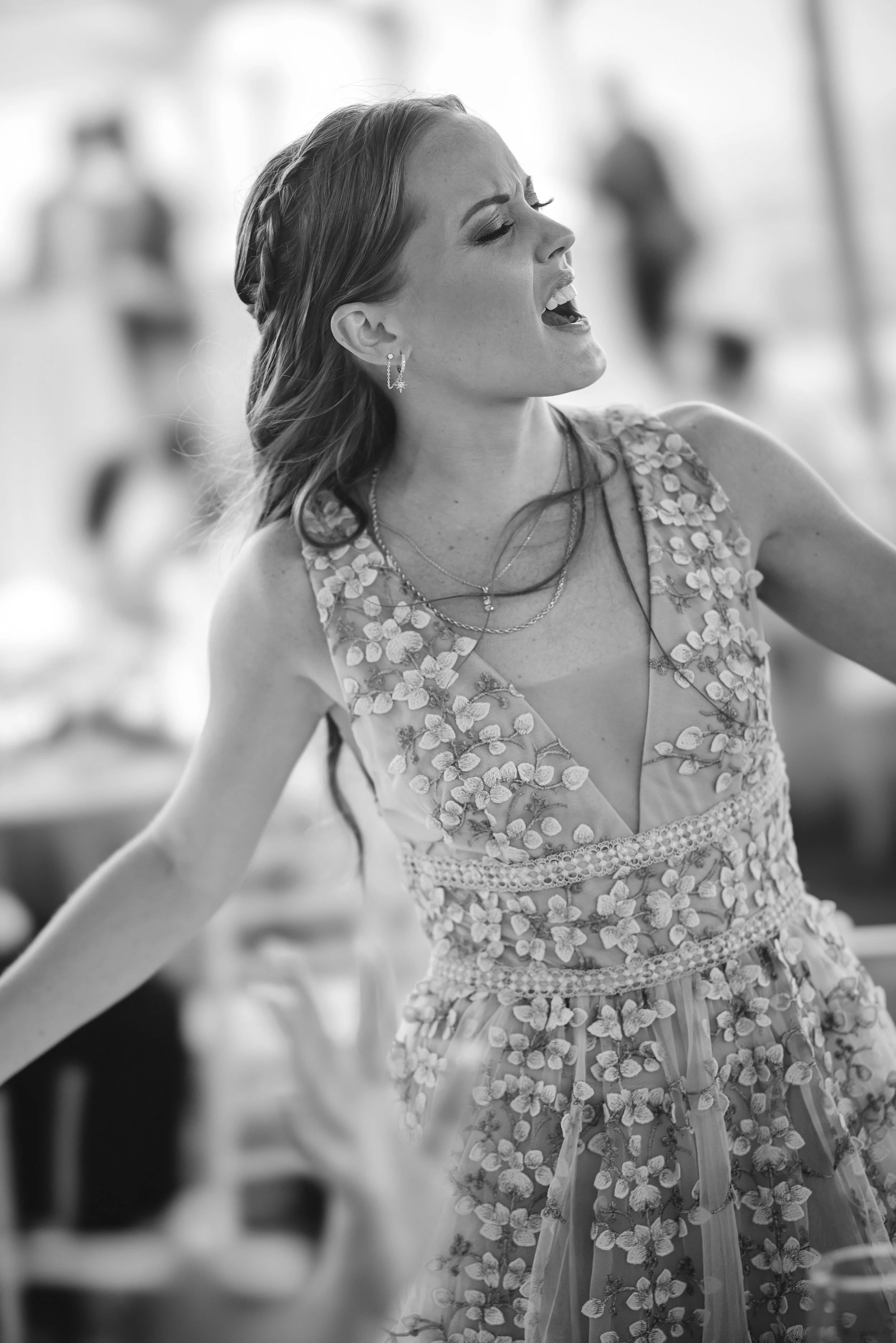 A woman in a floral dress sings passionately, head tilted back and mouth open, her expression full of emotion as she performs among blurred guests.