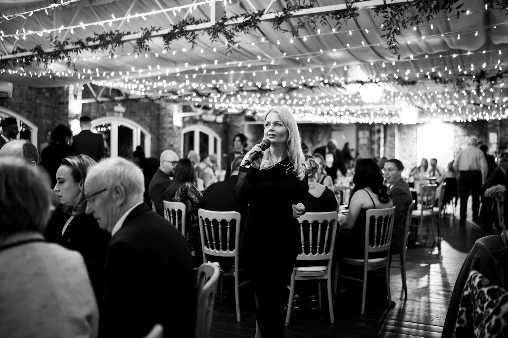A woman sings into a microphone among seated guests at a formal dinner, fairy lights overhead creating a glowing, festive atmosphere.