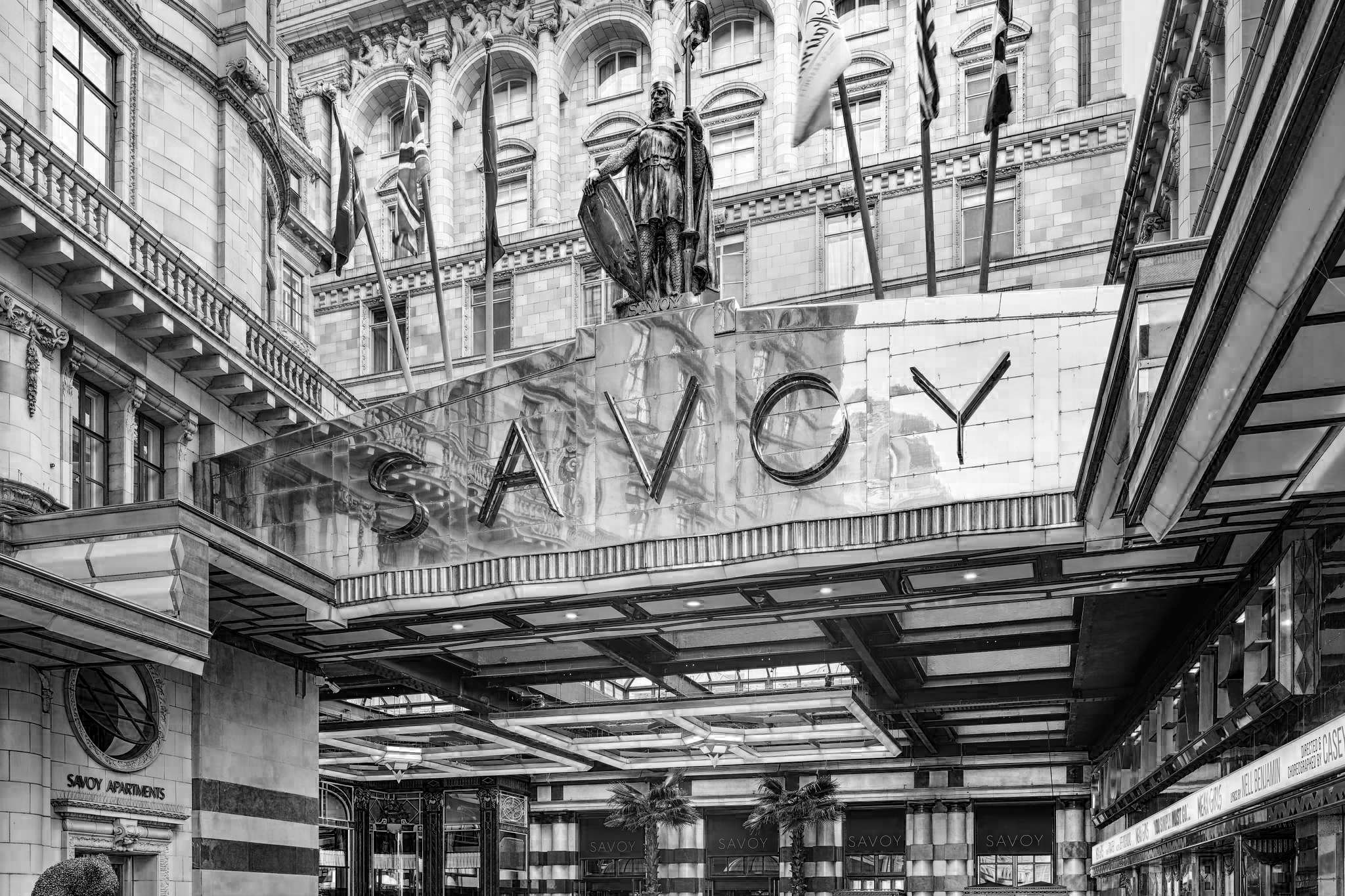 Black-and-white view of The Savoy hotel entrance in London, with a large Savoy sign, glass canopy, flags above, and a statue standing above the doorway.
