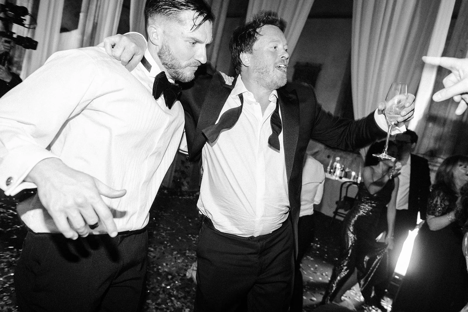 Two men in tuxedos dance arm in arm on a confetti-strewn floor, one raising a champagne glass as guests celebrate around them.
