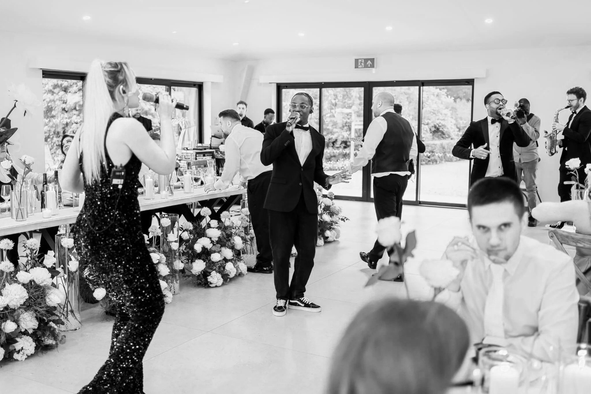 Two singers in suits and a sequined dress perform among wedding tables, flowers, and candles as guests watch in a bright, modern room.