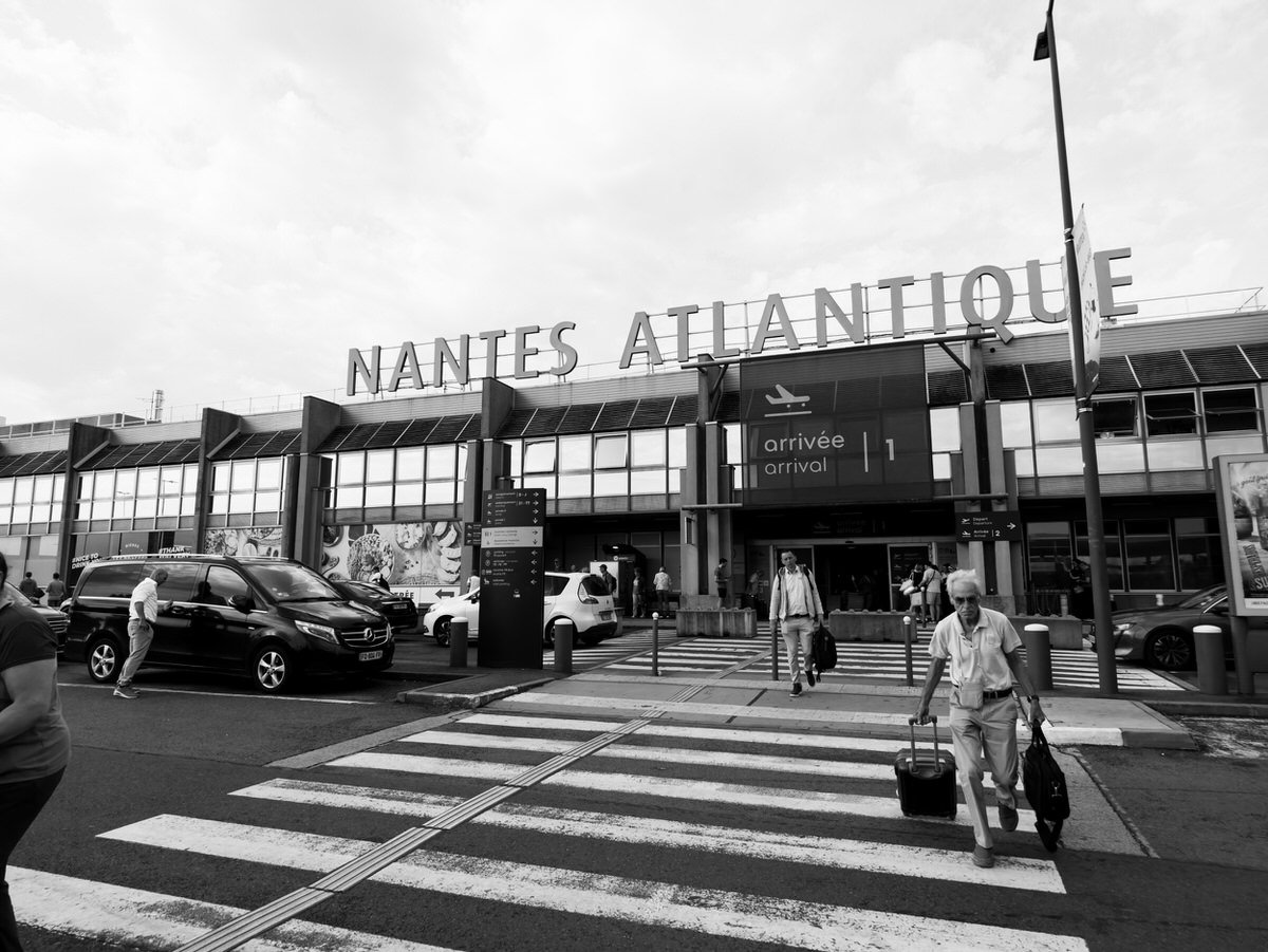 Exterior view of the Nantes Atlantique airport terminal entrance, a common destination for travelers, with people walking across a crosswalk.