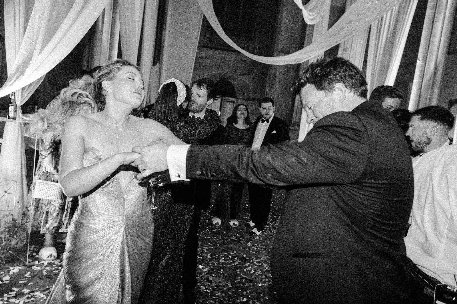 On a confetti-strewn dance floor, a woman in a strapless gown spins with a suited man, guests watching beneath flowing drapes.