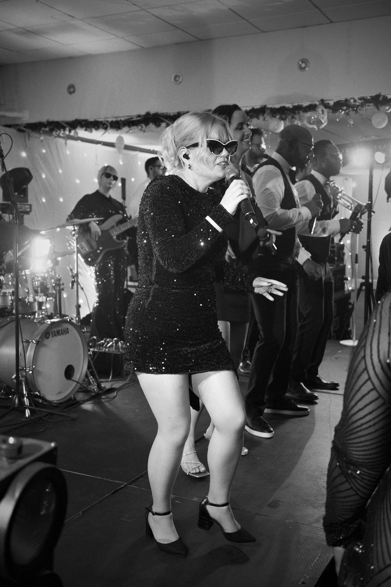 A singer in a sparkly short dress and sunglasses performs on stage with a full band behind her, lights glowing as musicians line up and play.