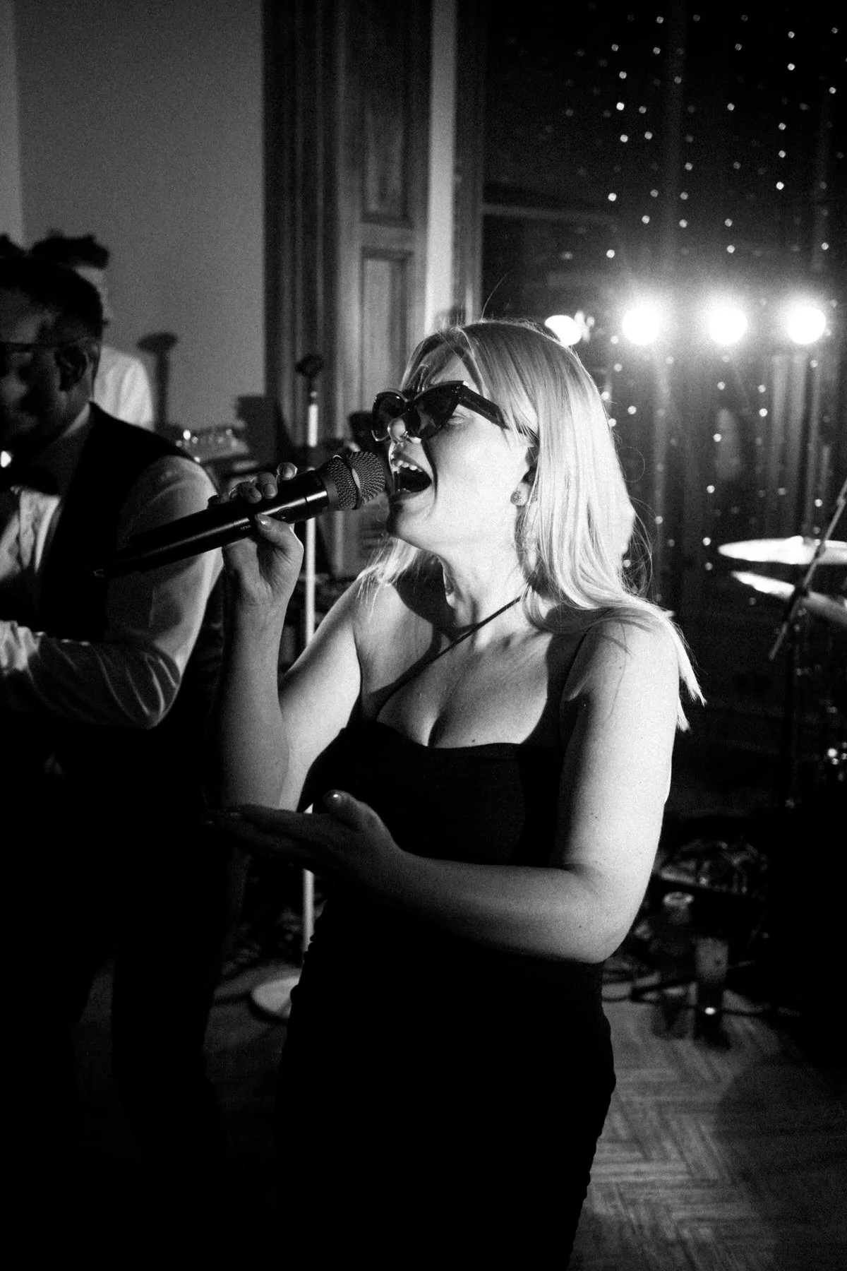 A black and white photo of a blonde woman wearing sunglasses singing into a microphone on stage, with a band member visible in the dark background.
