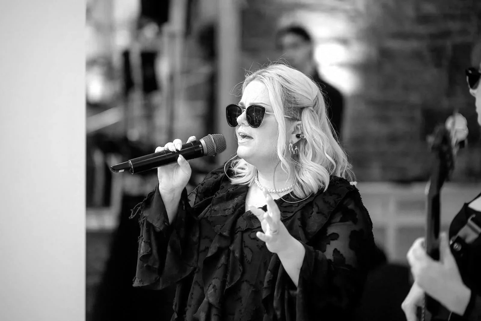 A blonde woman in a black dress and sunglasses sings into a microphone, one hand raised, pink lights glowing behind her.