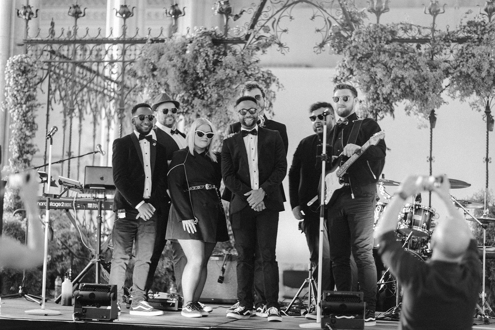 Seven smartly dressed band members in dark suits and sunglasses pose on a stage with guitars and drums, greenery arching behind them.