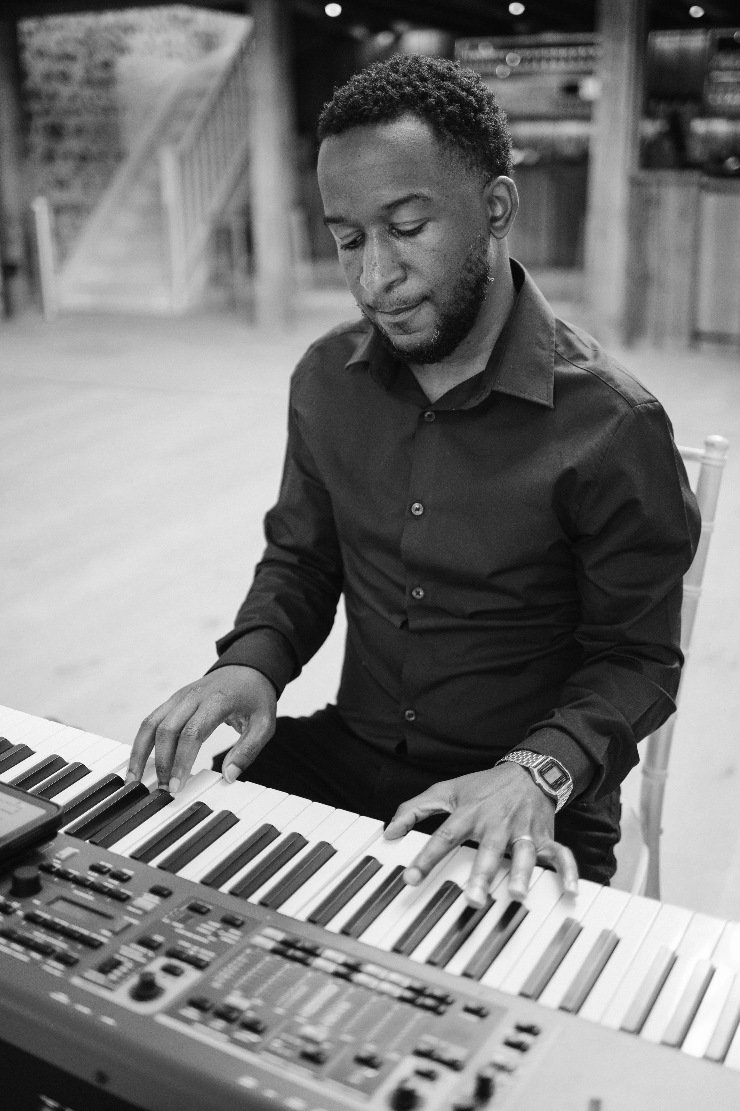 Black-and-white portrait of a man in a dark shirt playing a keyboard, focused, in a spacious room with stairs behind him.