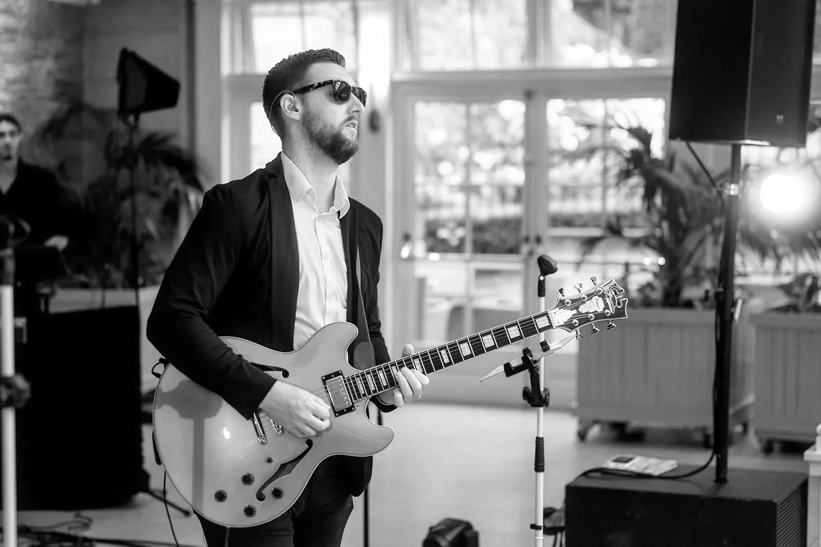 A man in sunglasses plays a mint-green electric guitar indoors, sunlight and greenery glowing through tall glass doors behind him.