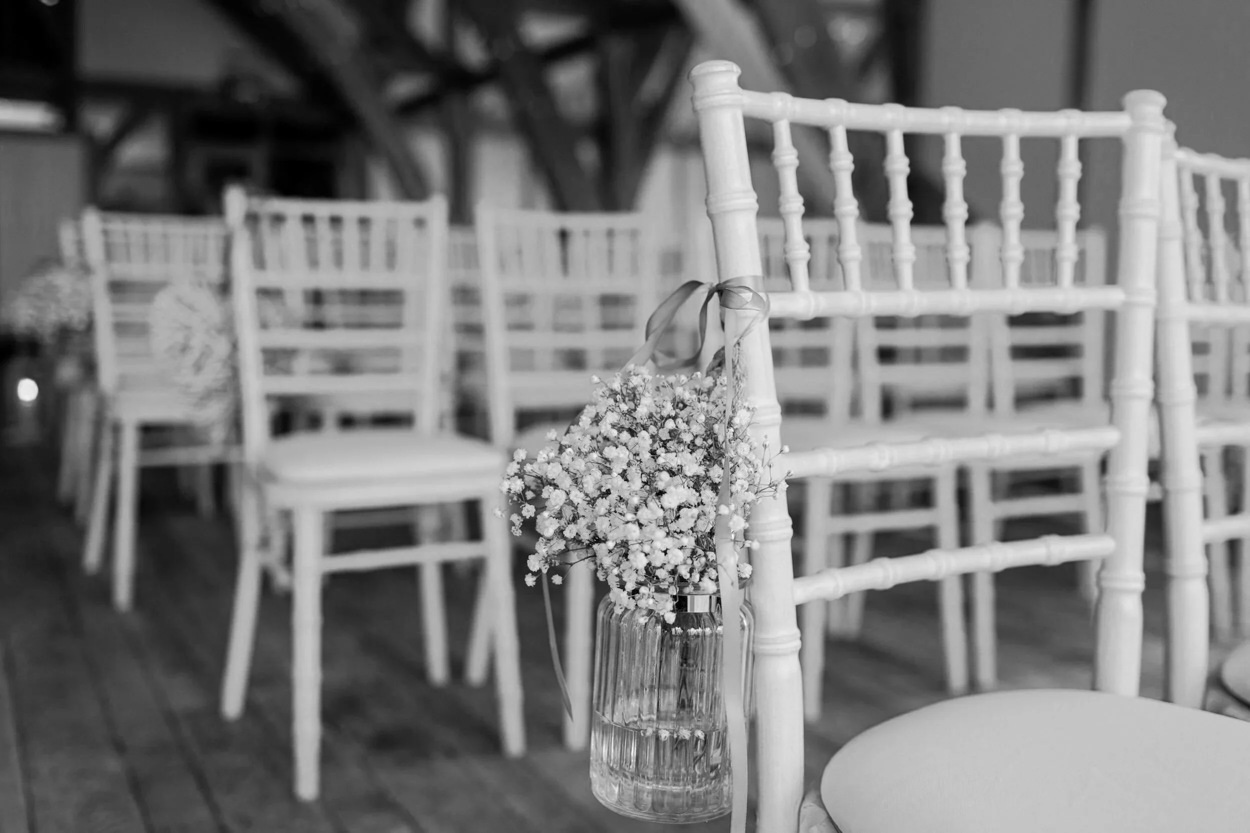 A glass jar of tiny white flowers hangs from a white chair with ribbon. Rows of matching chairs fade softly behind on a wooden floor.