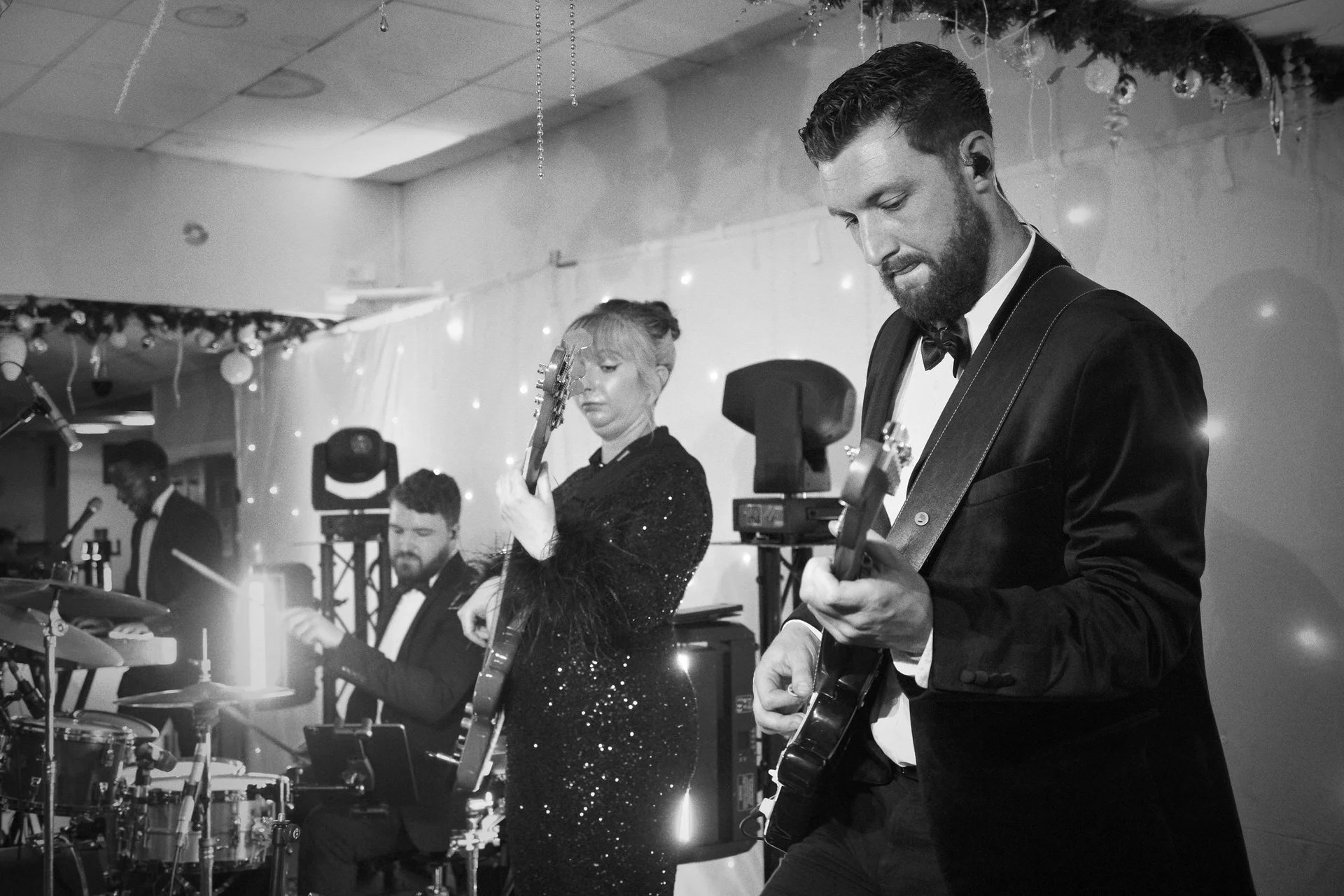 A formal band performs on stage: two guitarists and a drummer in tuxedos, fairy lights glowing behind them during an elegant event.