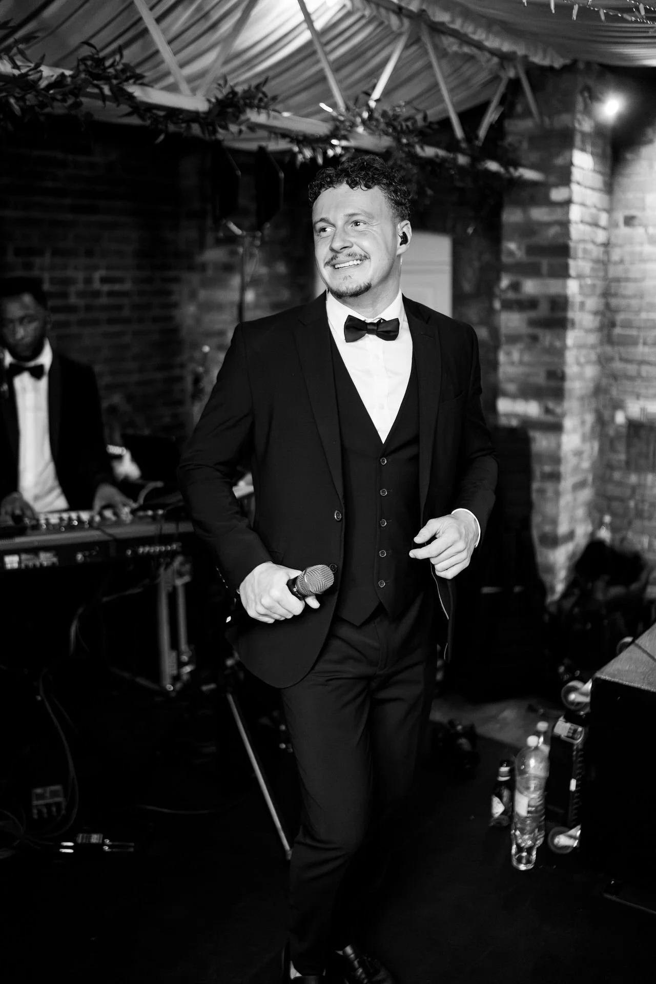 A smiling singer in a tux holds a microphone on a small stage, brick walls and draped lights behind, with a keyboardist blurred in the background.