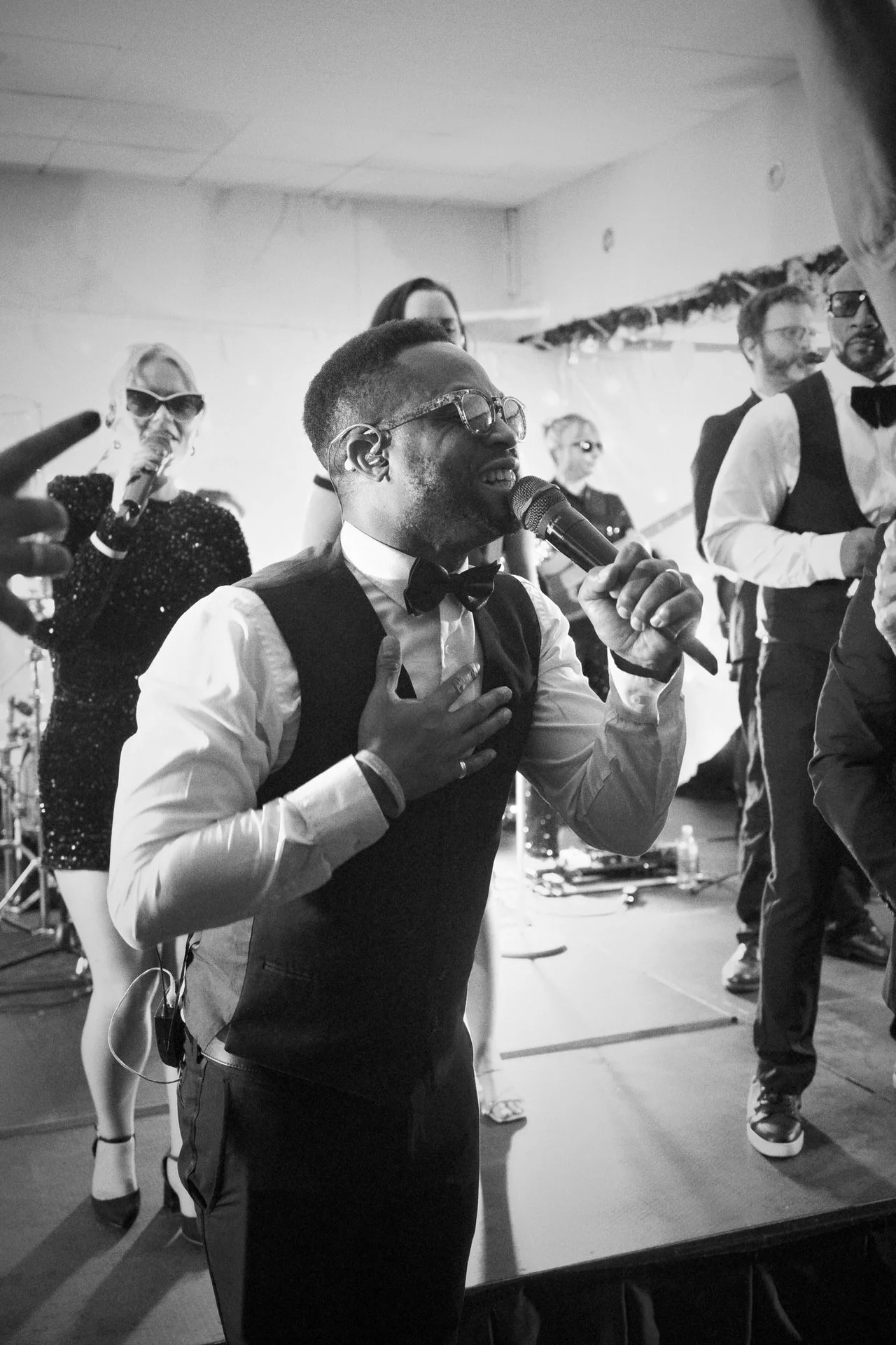 A smiling singer in a vest and bow tie holds a mic to his mouth, hand on his chest, while bandmates perform behind him on a bright stage.