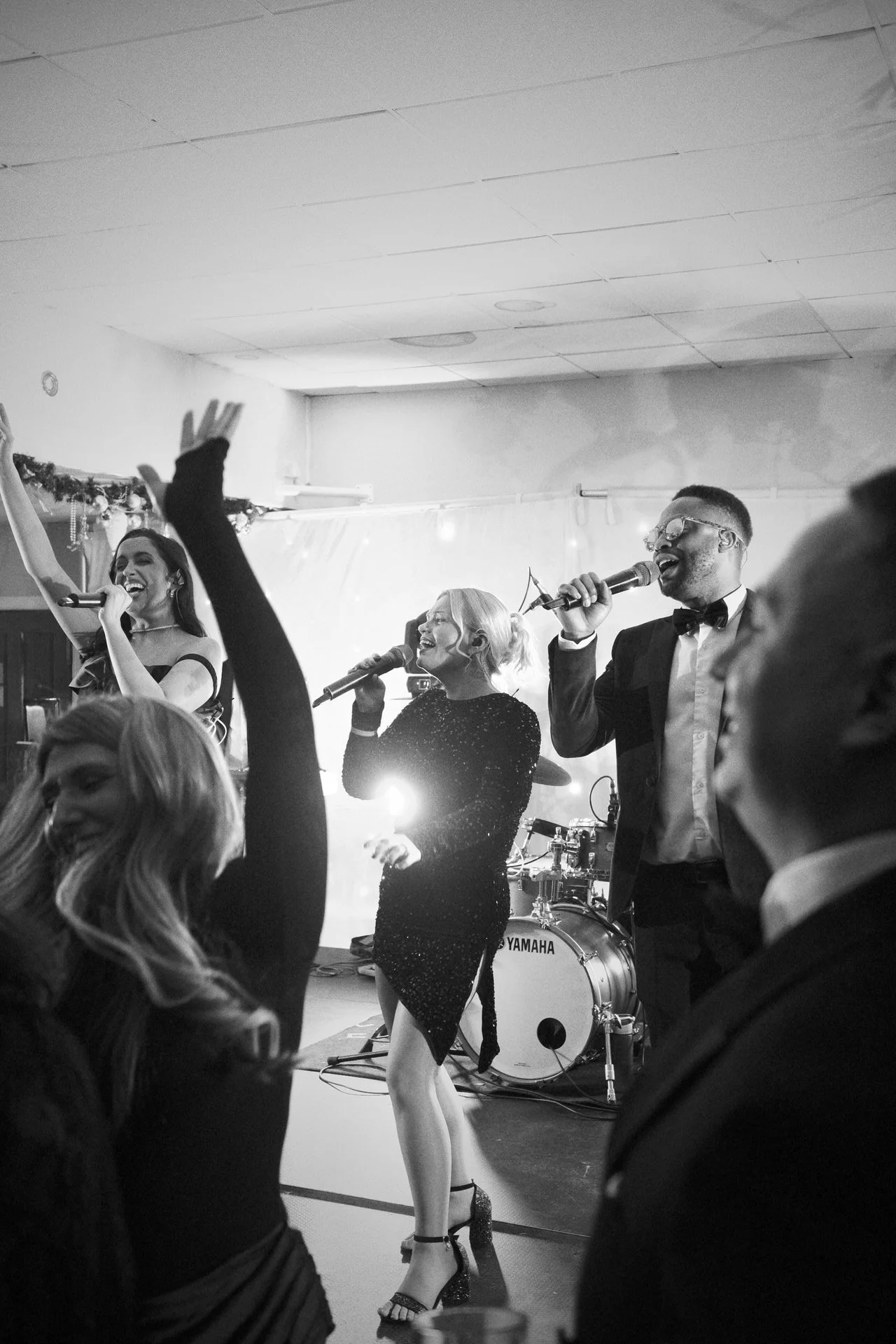 Three singers in formal wear perform on stage as guests raise their arms dancing; bright lights and drums glow behind them in a lively celebration.