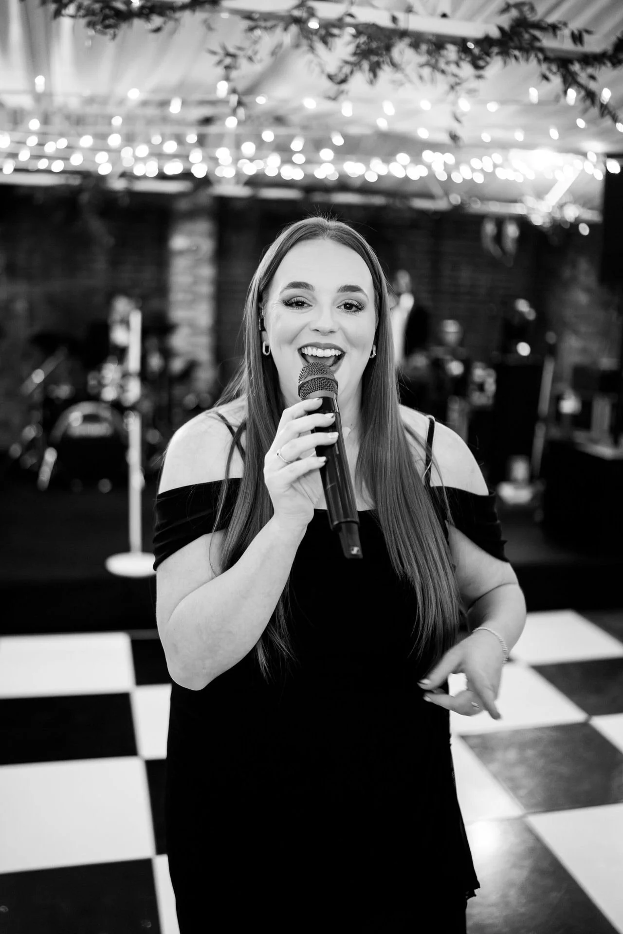 A smiling singer in a long dress holds a microphone on a checkered dance floor, with fairy lights and a blurred band behind her.