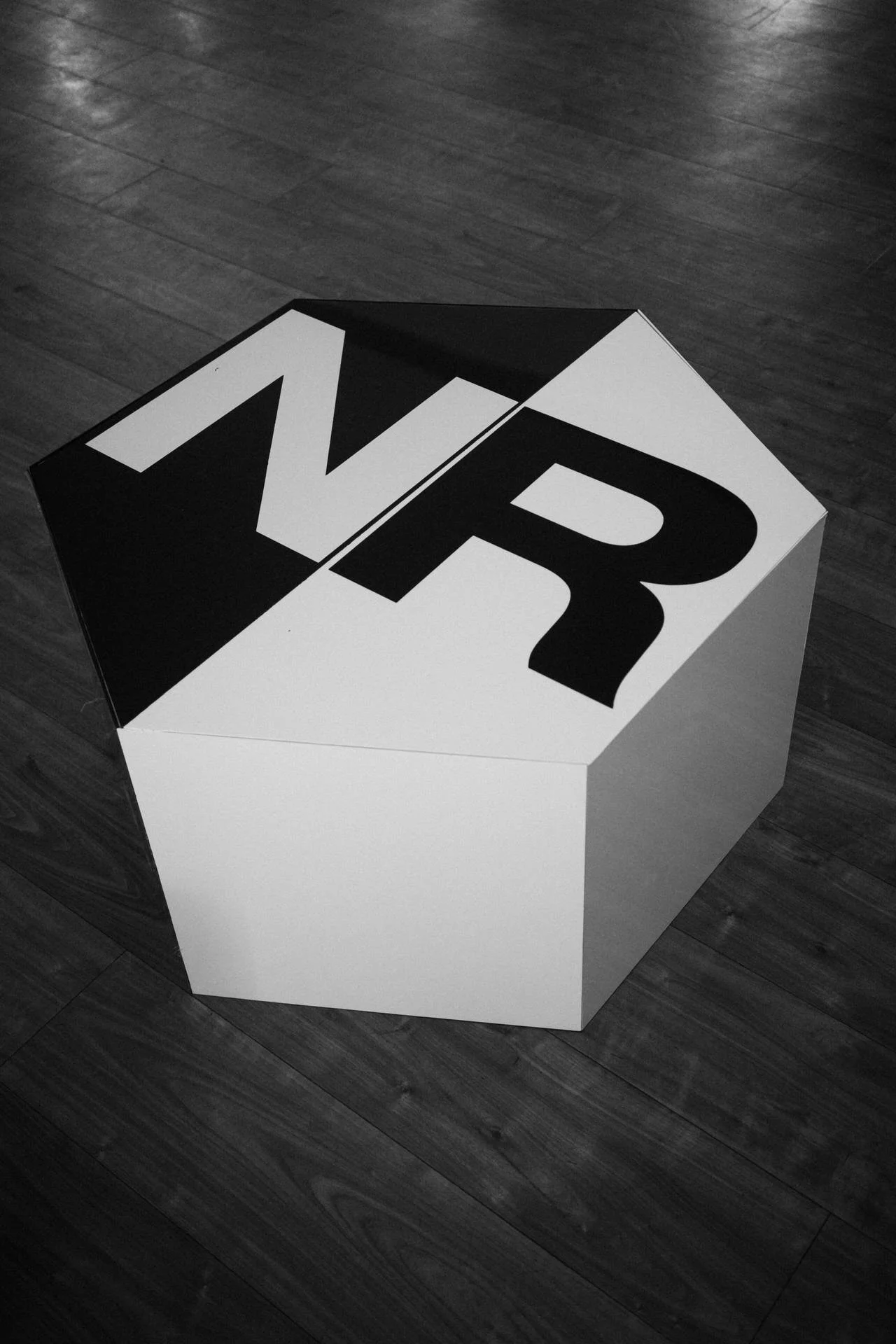 A black-and-white photo of a white cube on a wooden floor, its top marked with bold black letters forming an abstract logo.
