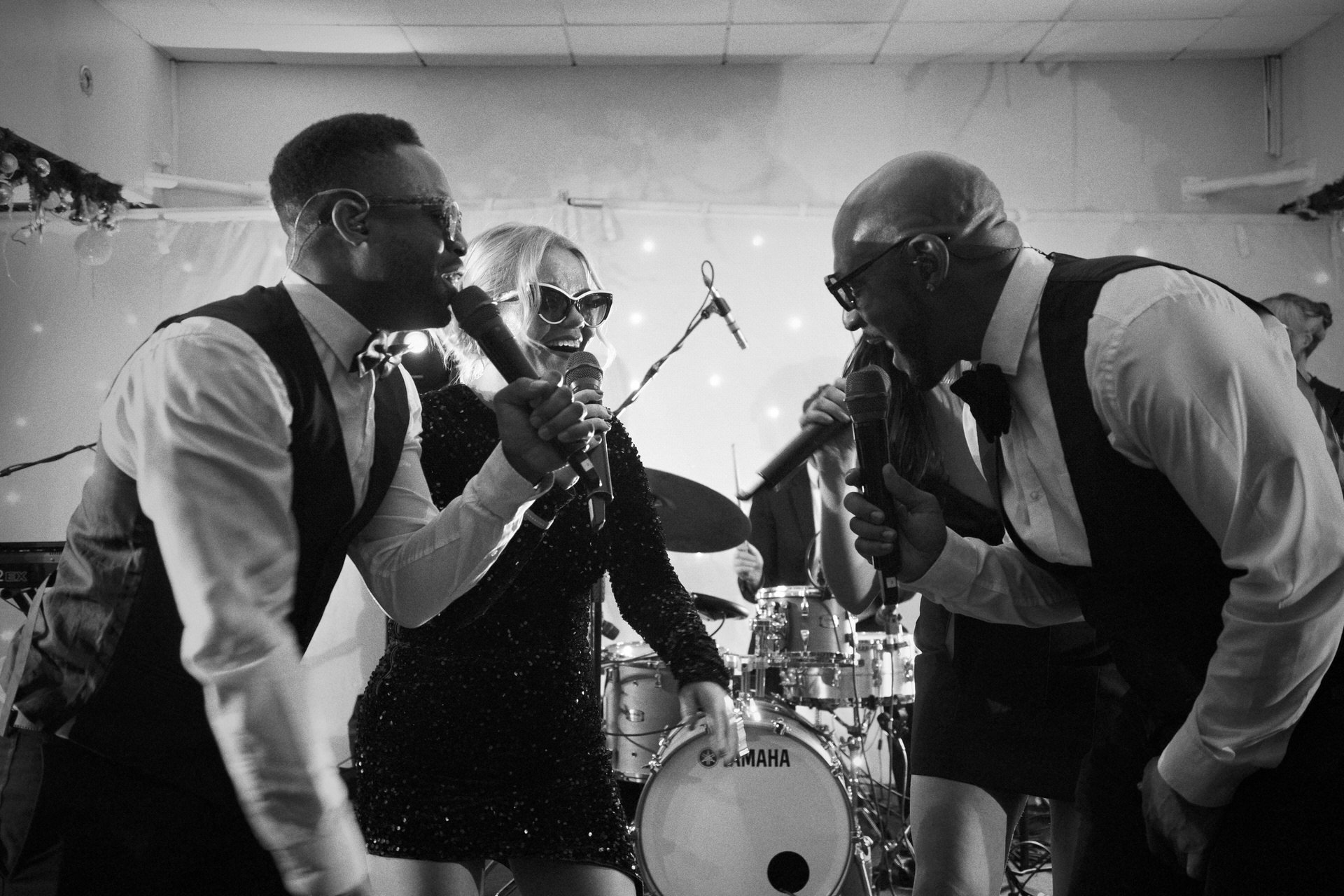 Three singers lean toward each other with microphones, smiling mid-song, while a drummer plays behind them under soft stage lights.