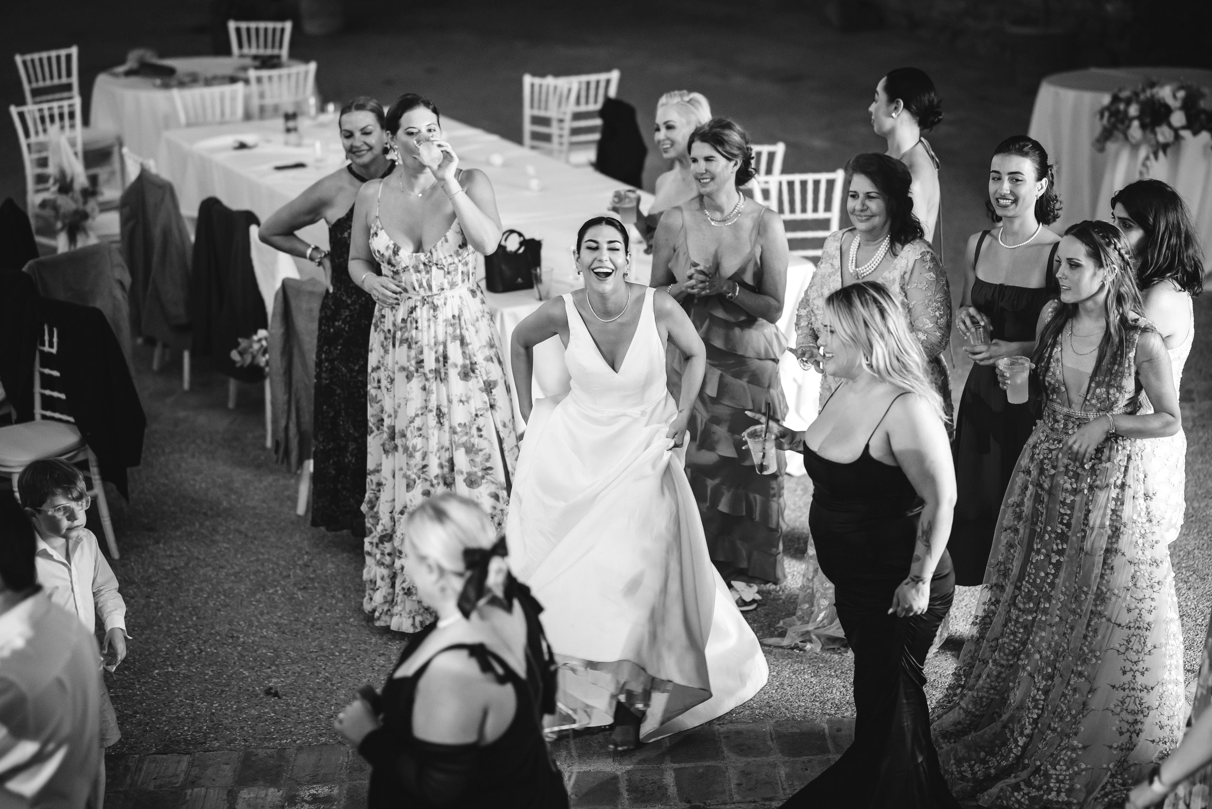 A joyful bride runs forward laughing as a group of women in formal dresses cheer and watch, drinks in hand, at a lively wedding reception.