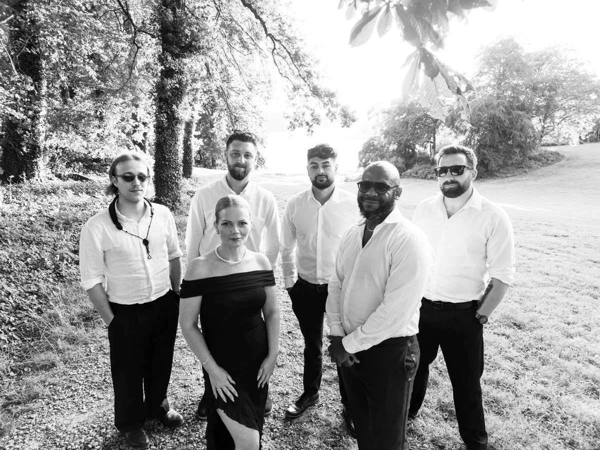 Black-and-white outdoor group portrait of six adults in formal attire beneath leafy trees, standing together by a sunlit path with a relaxed, celebratory mood.