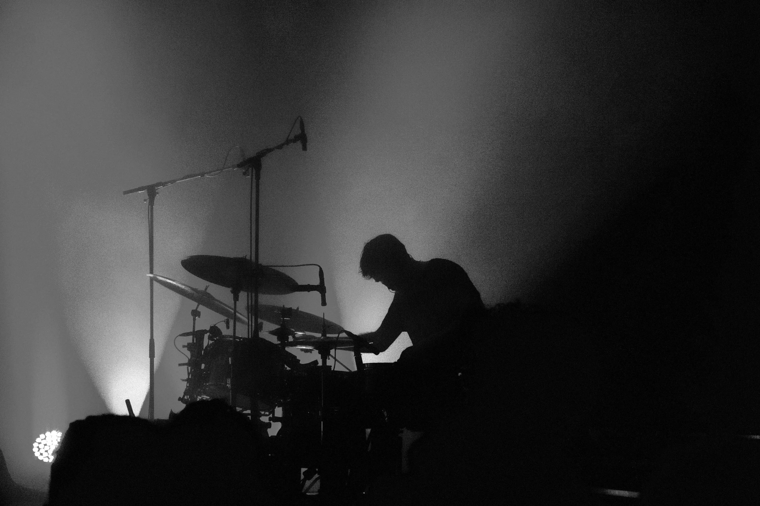 A drummer’s silhouette plays onstage, framed by blue and pink beams of light, with cymbals and microphones glowing in smoky haze.