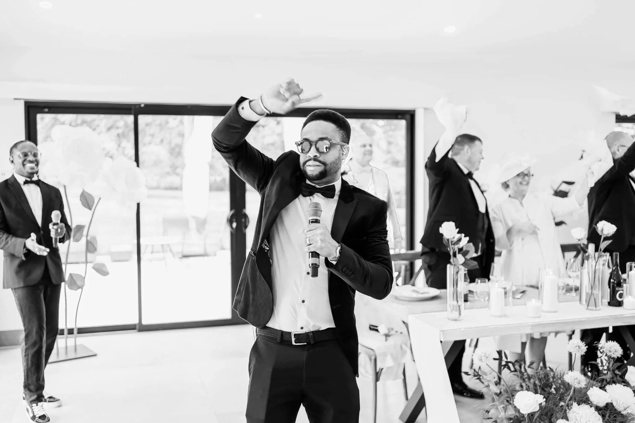 A tuxedoed singer raises one arm and holds a mic as guests cheer at tables behind him, waving napkins in a bright, flower-filled reception room.