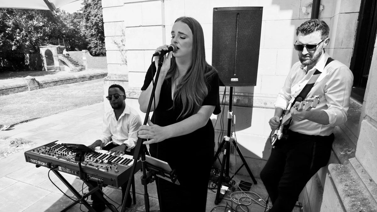 A black and white photo of a musical trio performing outdoors, featuring a female singer, a keyboardist, and a guitarist standing near a building.