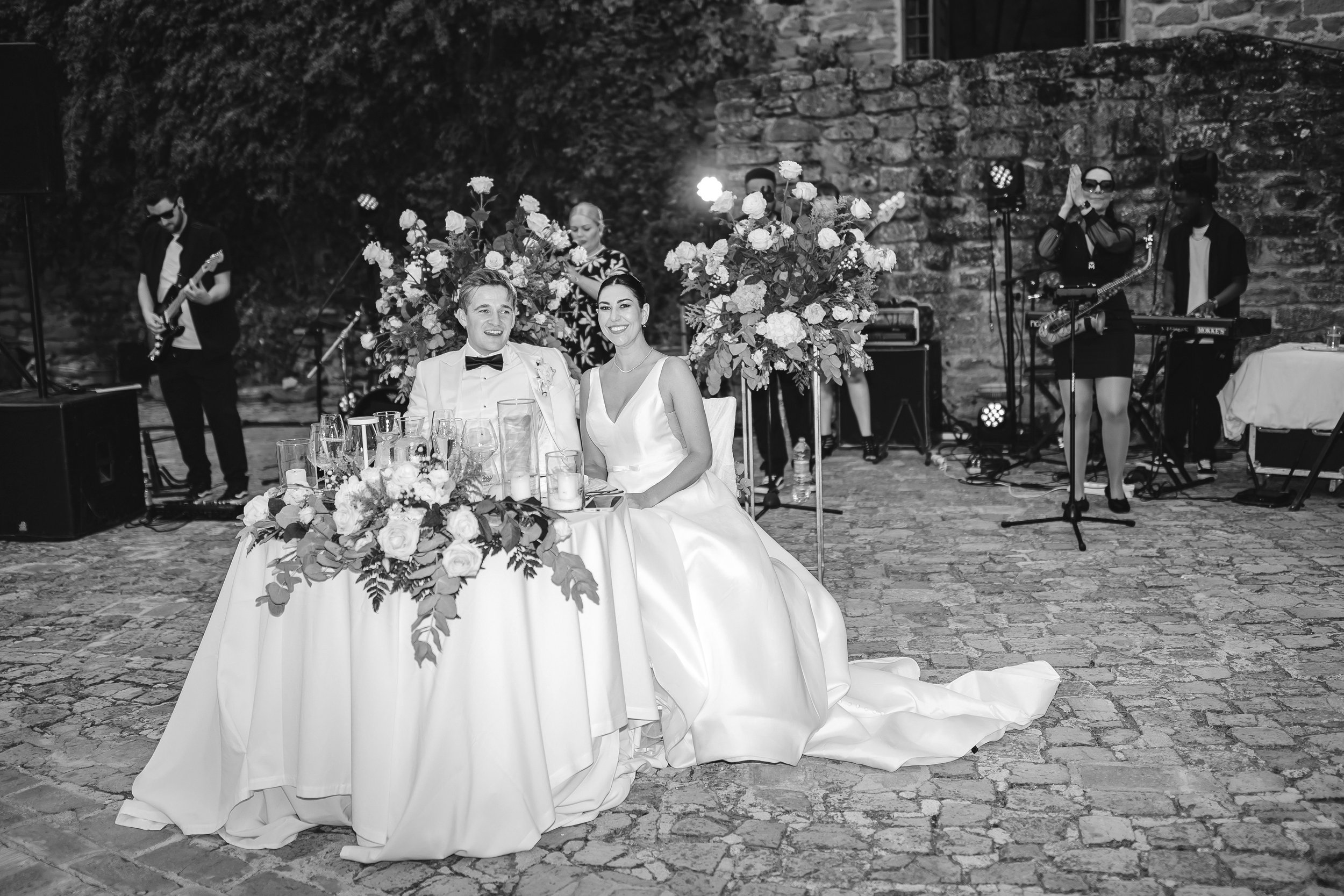 A bride and groom sit smiling at a flower-covered table in a stone courtyard while a live band performs behind them under soft lights.
