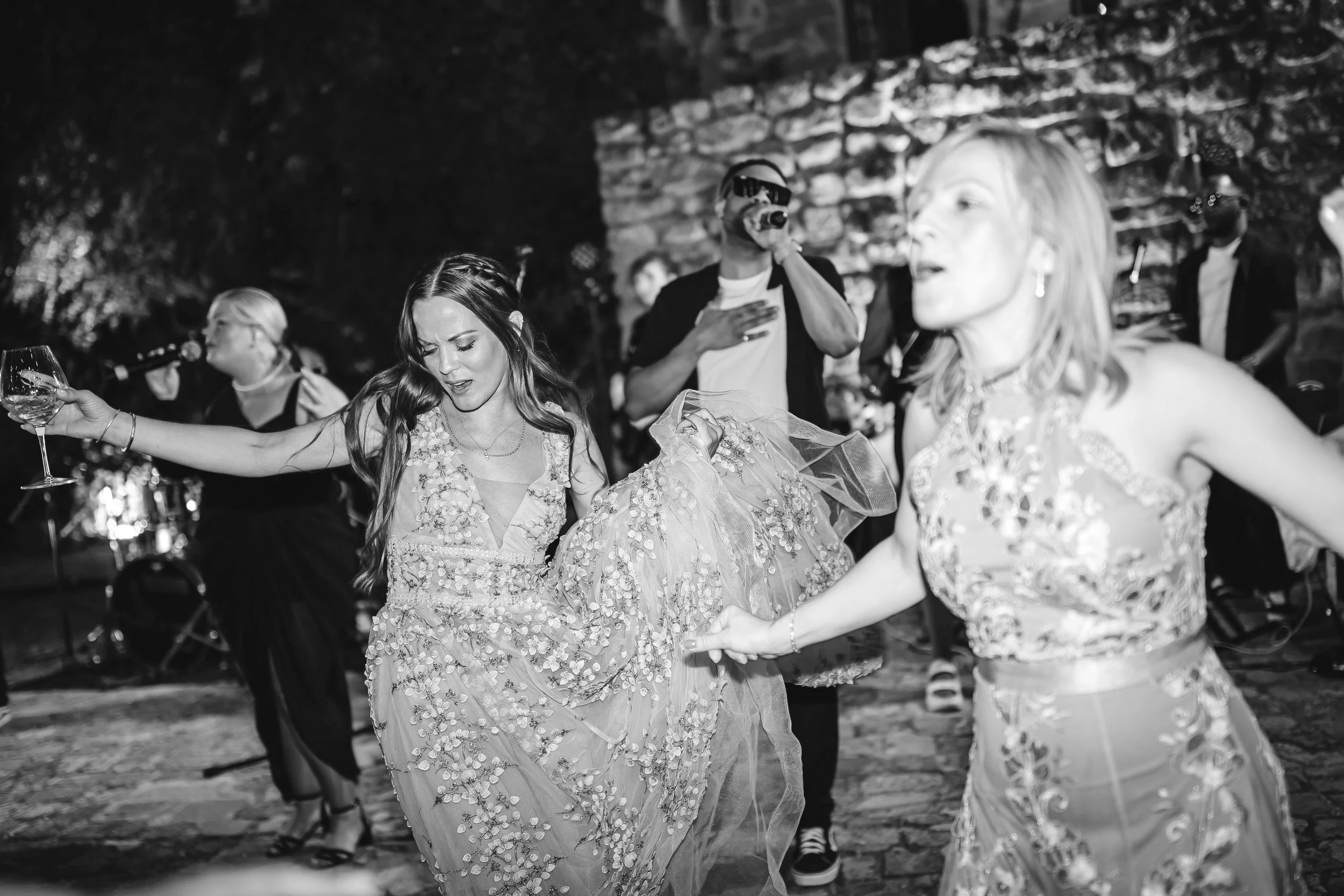 Two women in flowing dresses dance with joy, one holding a wine glass, while a singer performs behind them in a lively outdoor celebration.