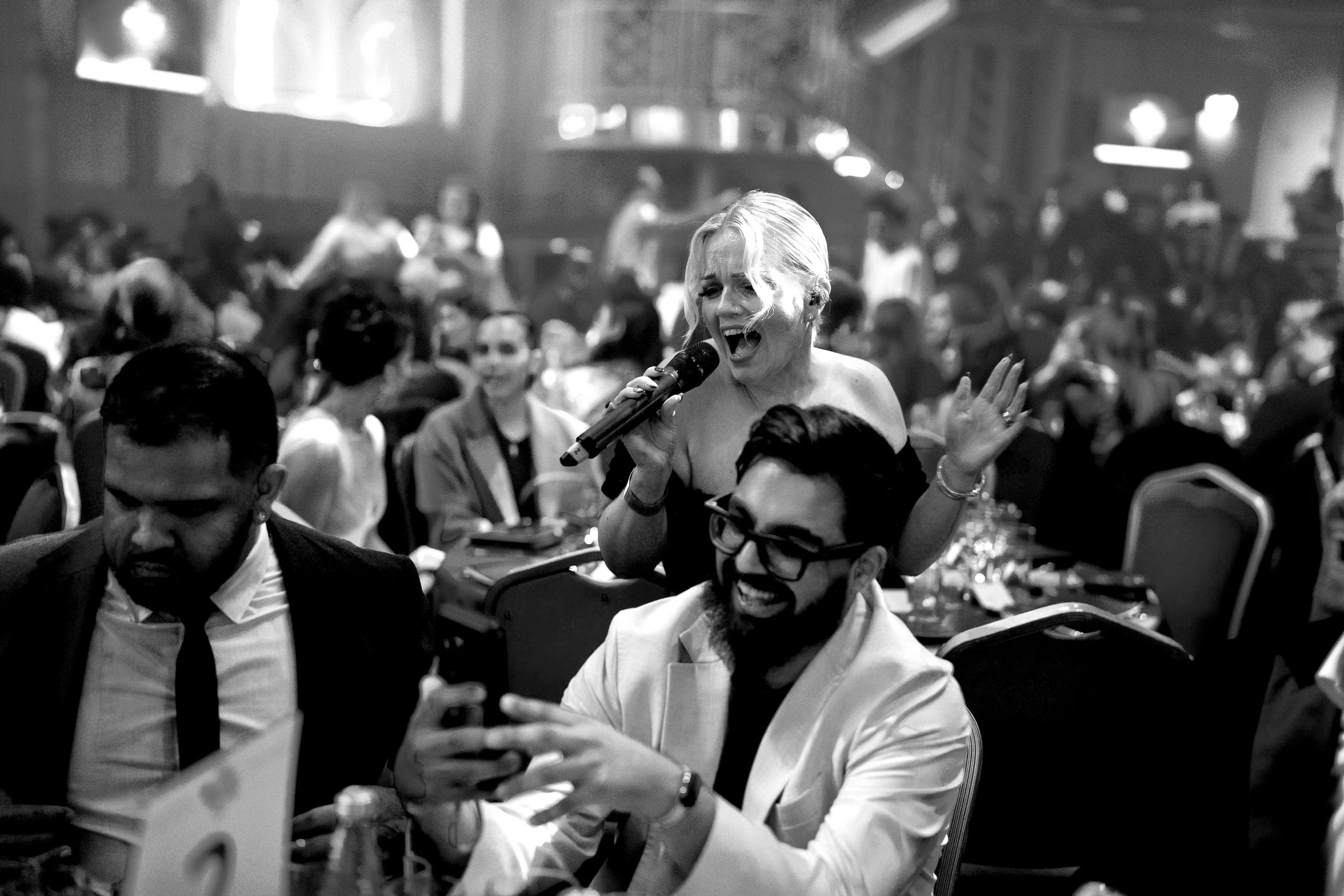 A singer performs among guests at a lively event, holding a microphone and singing beside tables while smiling attendees watch and film the moment.