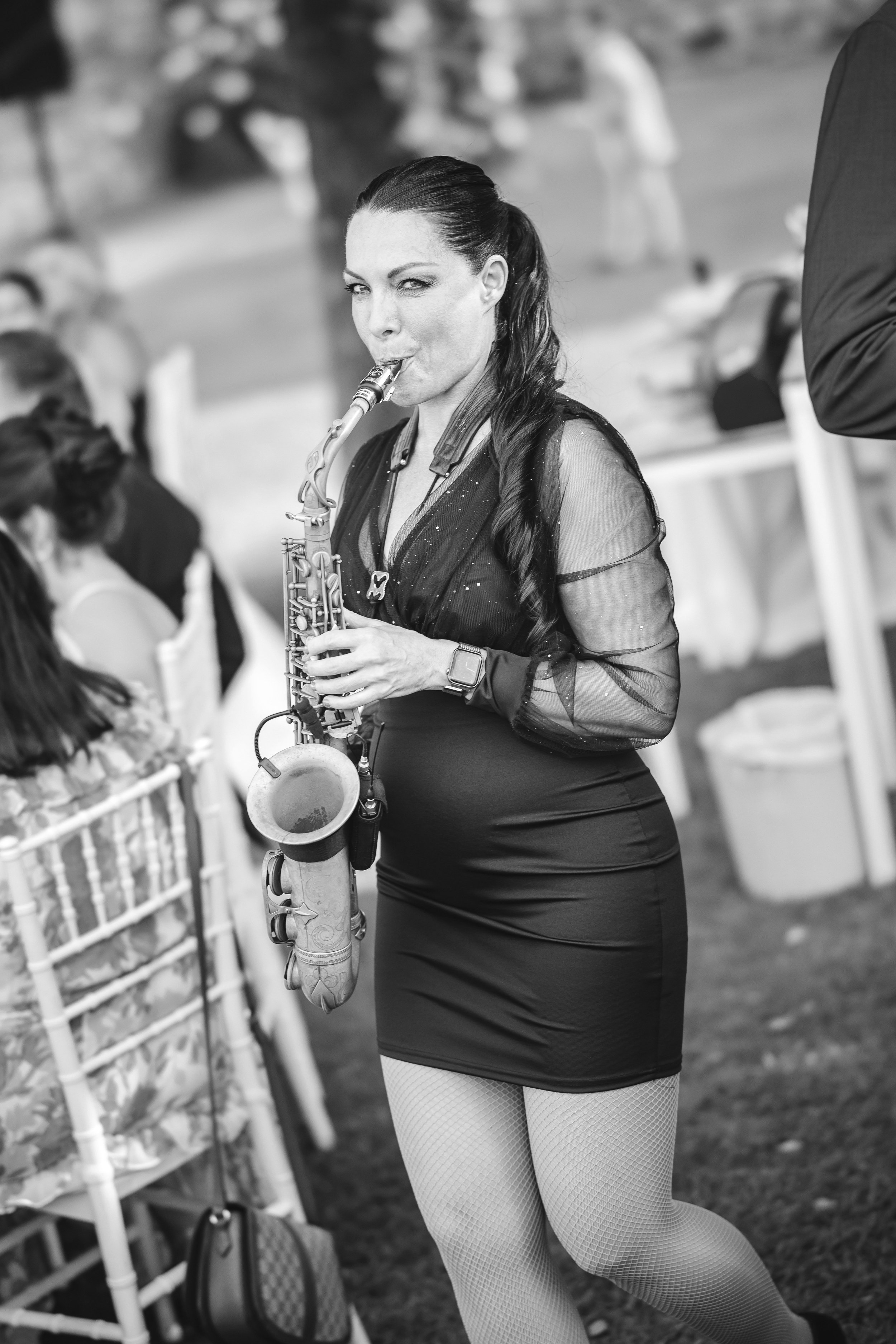A woman in a fitted dress walks among guests, playing a saxophone, her posture confident as people sit nearby listening in an outdoor setting.