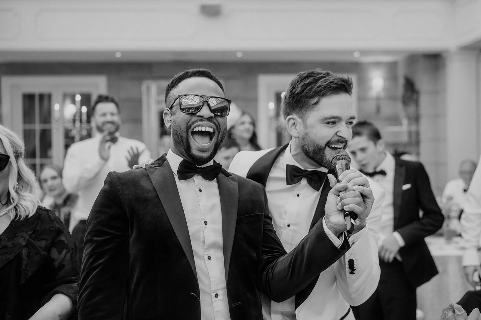 Two men in tuxedos grin and sing into a microphone, one in sunglasses, as wedding guests cheer behind them.