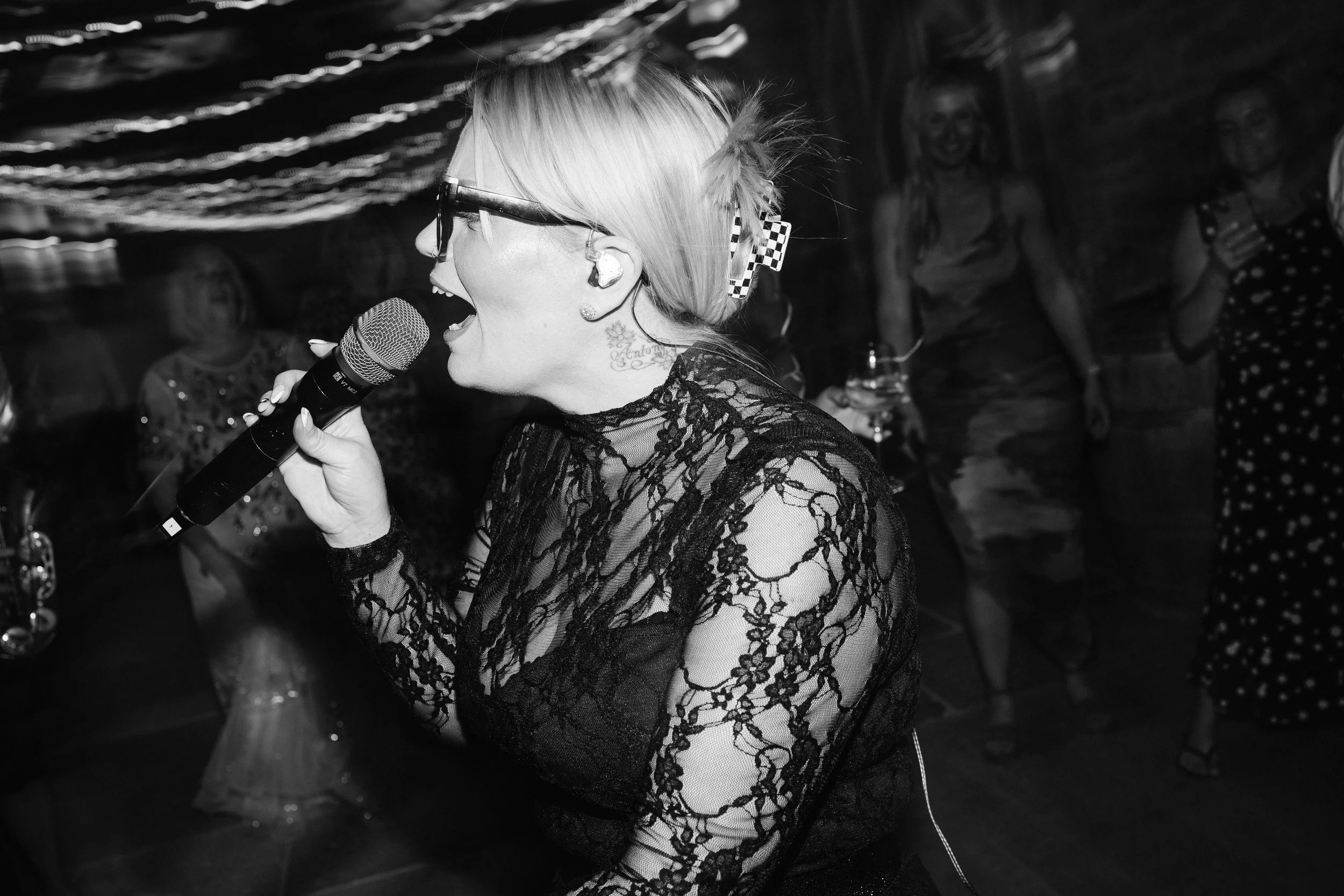 Black-and-white close-up of a singer in lace, mouth open mid-song, holding a mic as guests smile under string lights.