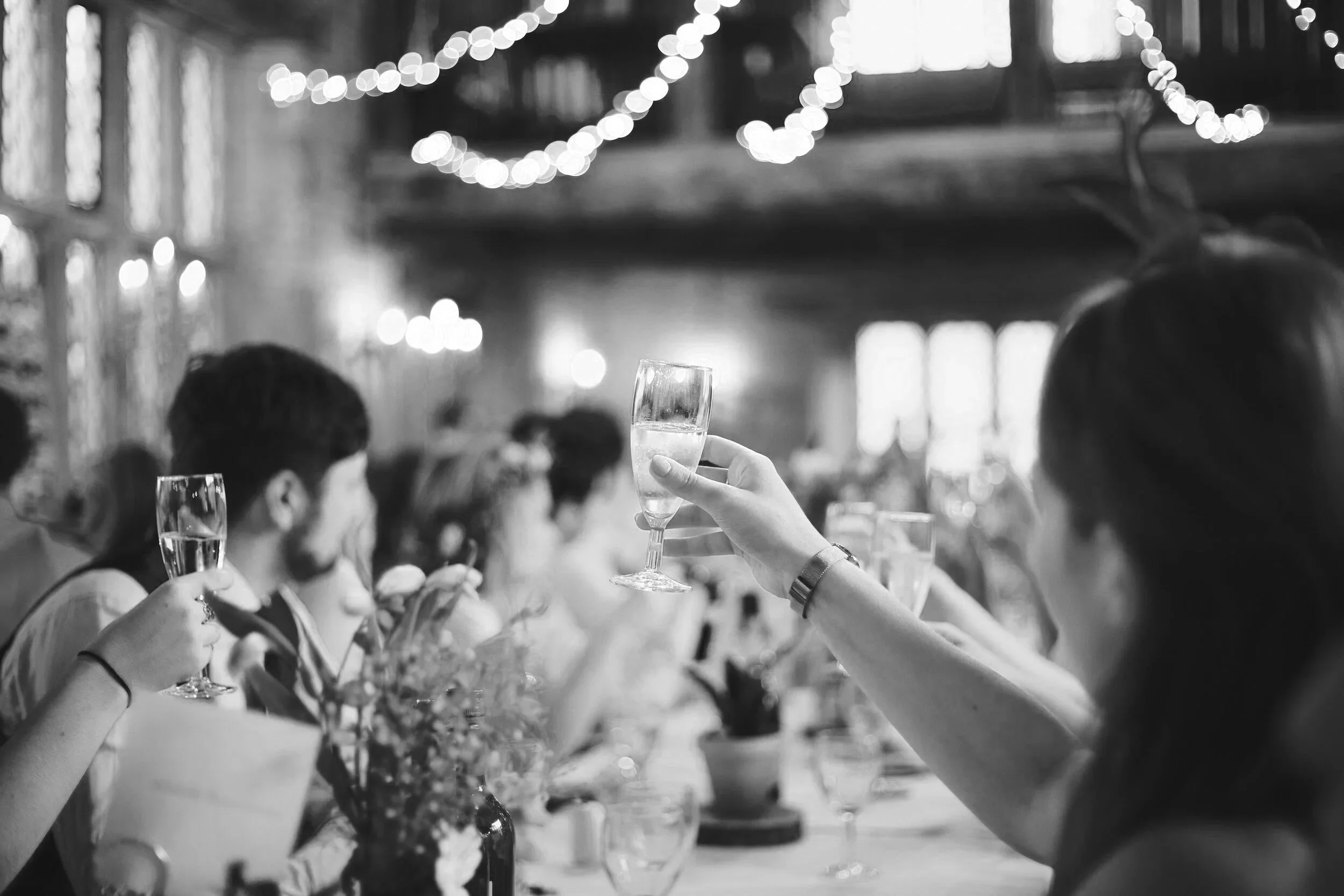 Guests seated at a long table raise champagne glasses in a toast. Soft lights glow overhead, creating a warm, festive atmosphere.