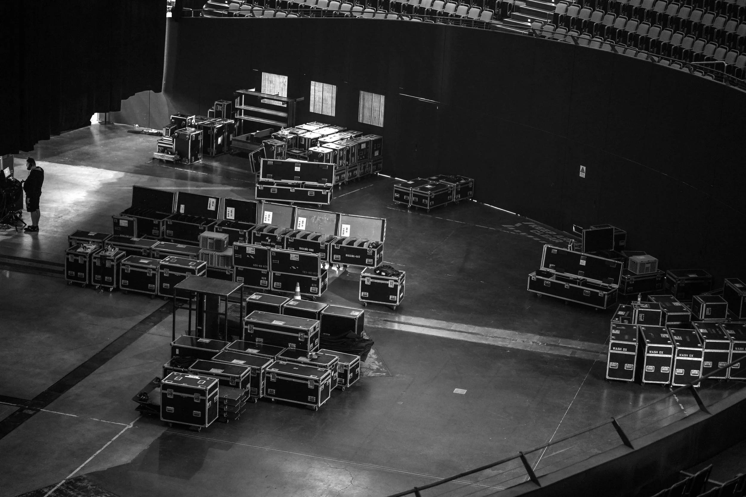 A wide arena floor filled with stacked black road cases and equipment, empty seats above, and a lone crew member working near the stage.