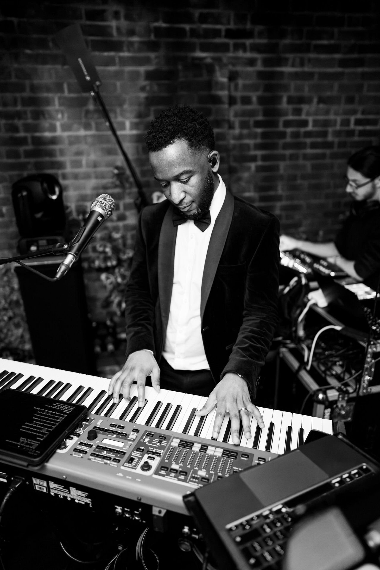 A tuxedoed keyboard player looks down at the keys mid-performance, a microphone nearby and brick walls behind him on stage.