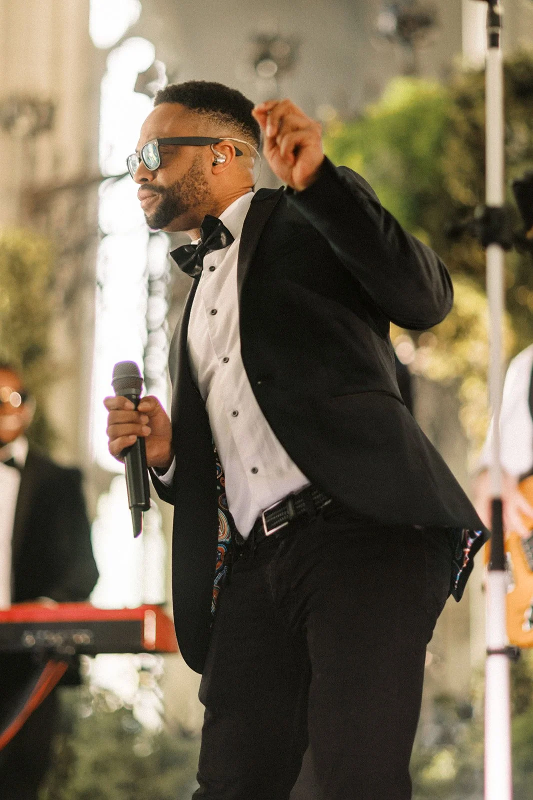 A man in a tuxedo and sunglasses sings into a microphone, mid-motion, performing energetically on an outdoor stage.