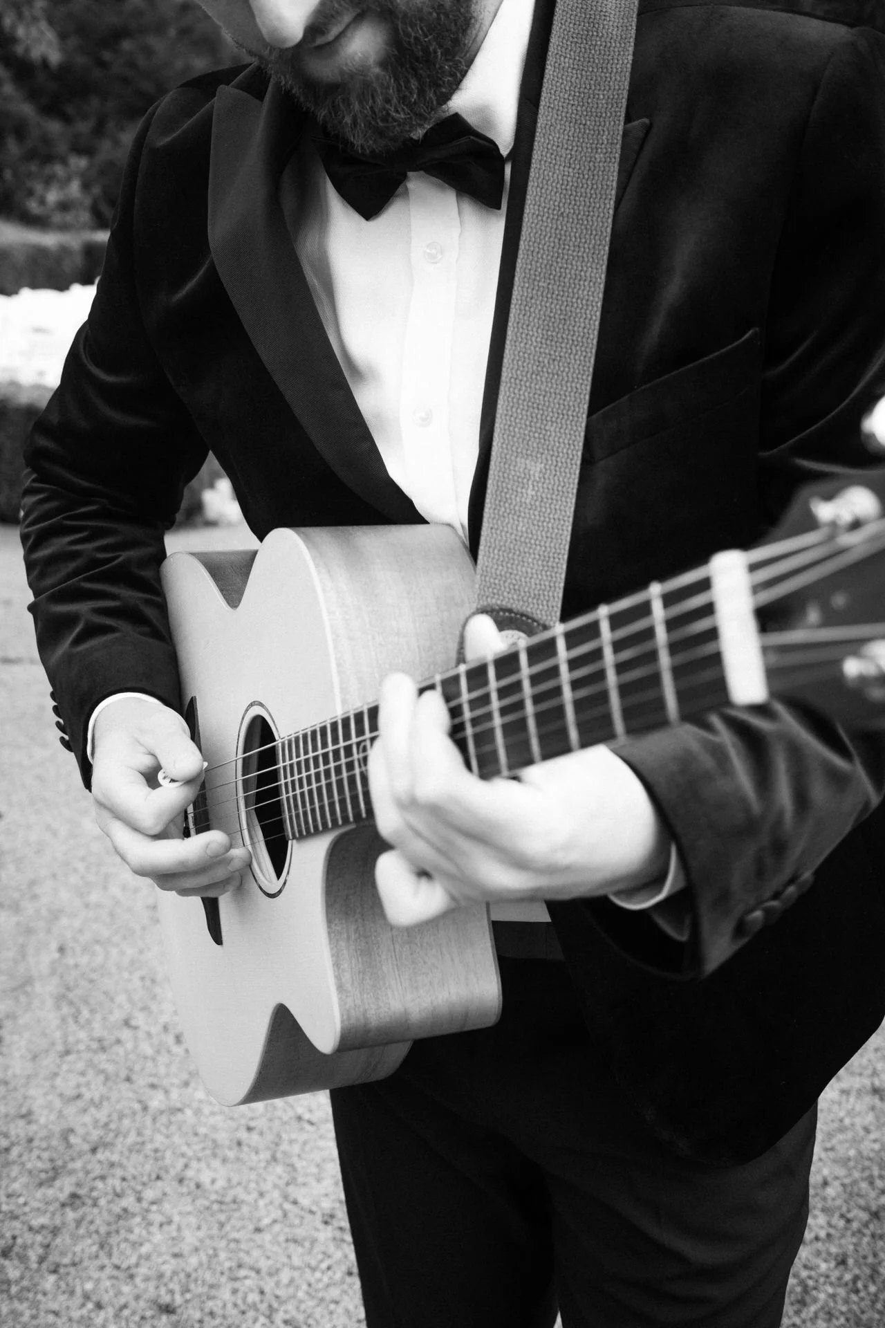 Close-up of a bearded man in a tuxedo strumming an acoustic guitar, his hands mid-chord, outdoors on a gravel path.