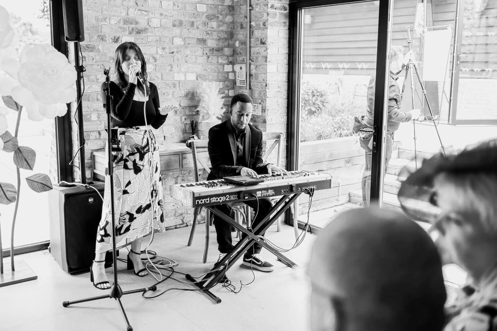 A woman sings into a mic while a man plays keyboard in a brick-walled room; guests watch as daylight streams through open glass doors.