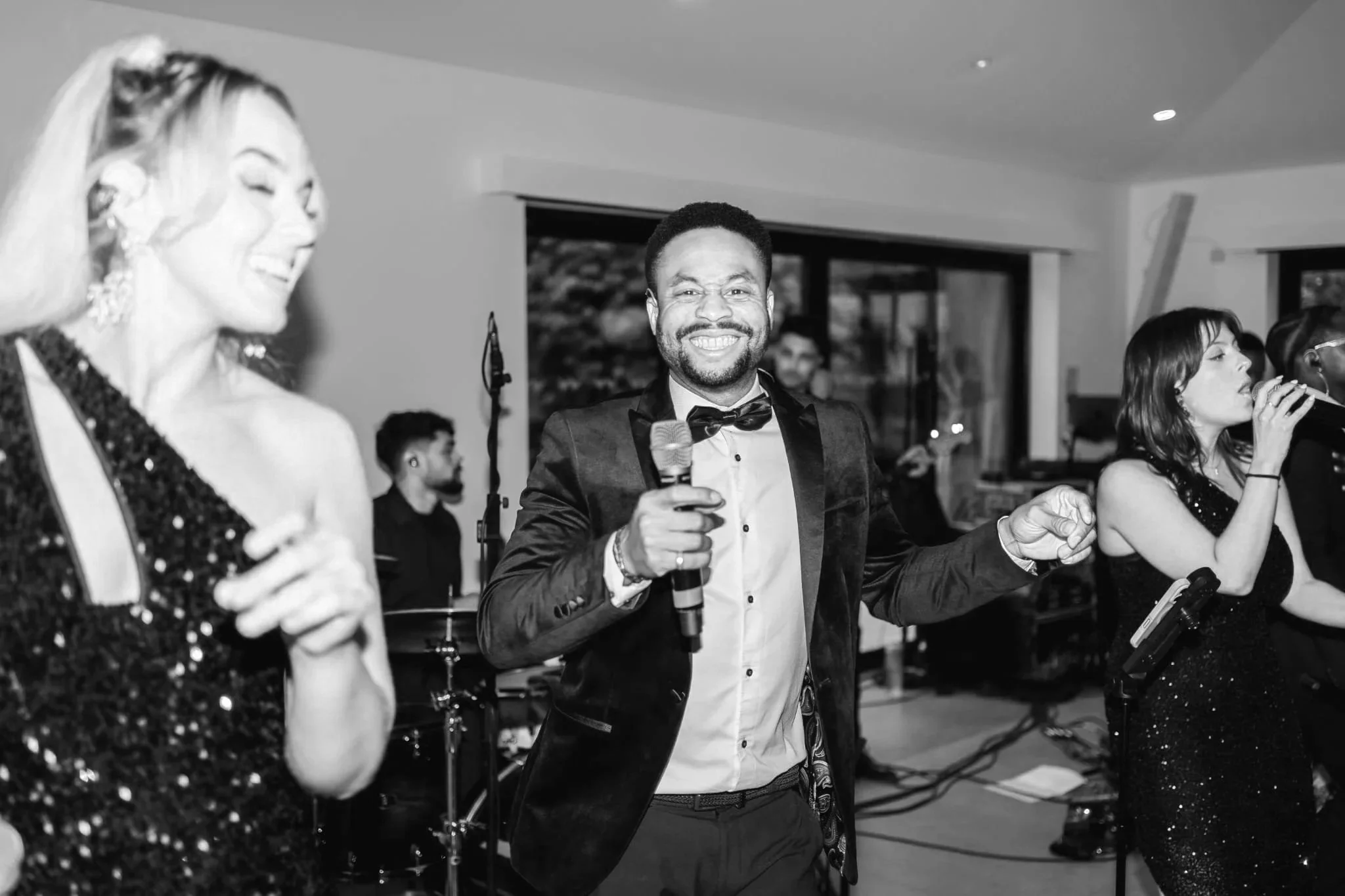 A smiling lead singer in a tux holds a mic between two glitter-clad vocalists, with a drummer and band playing behind them.