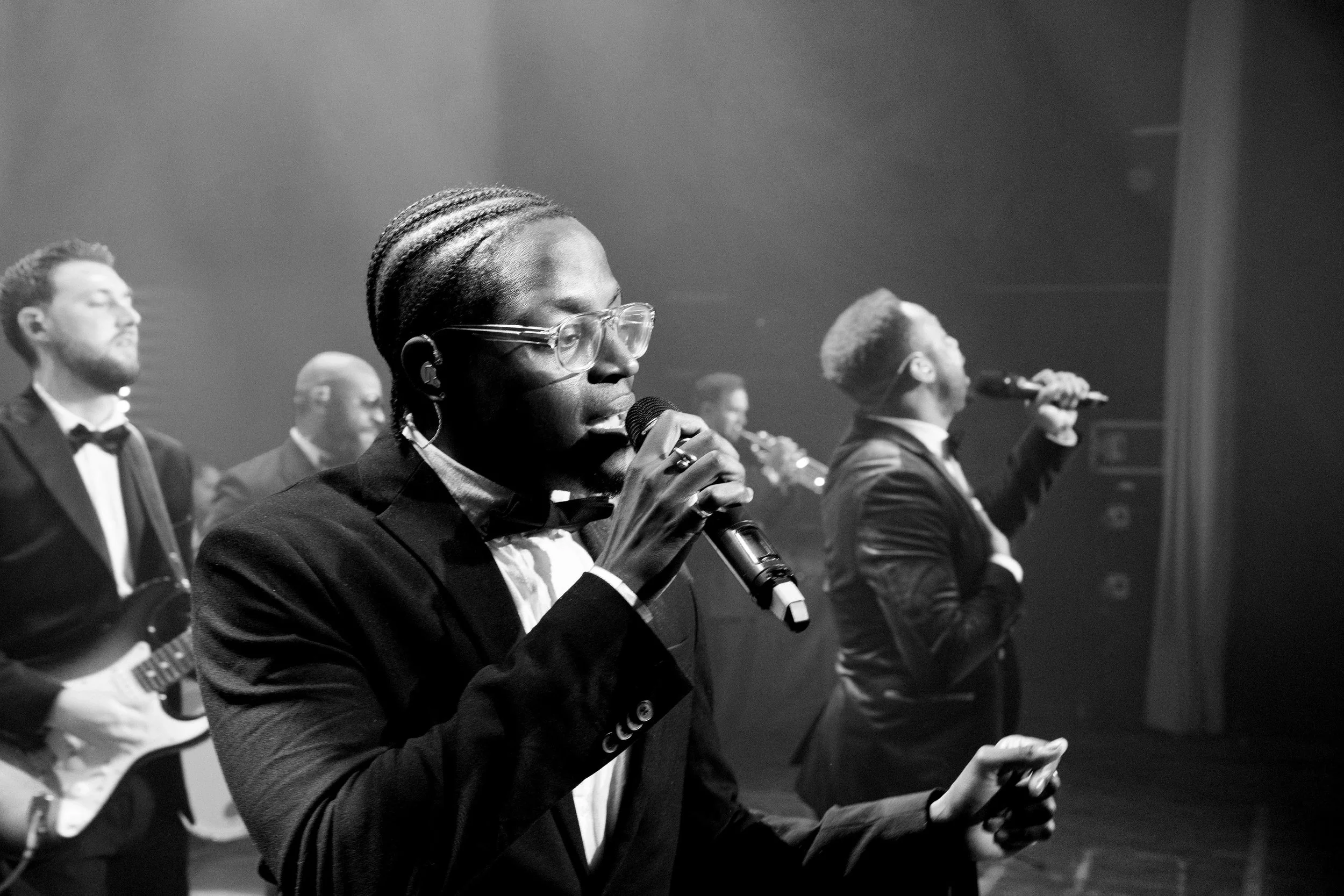 A sharply dressed singer performs with a live band on stage, holding a microphone as fellow musicians and vocalists play behind him.